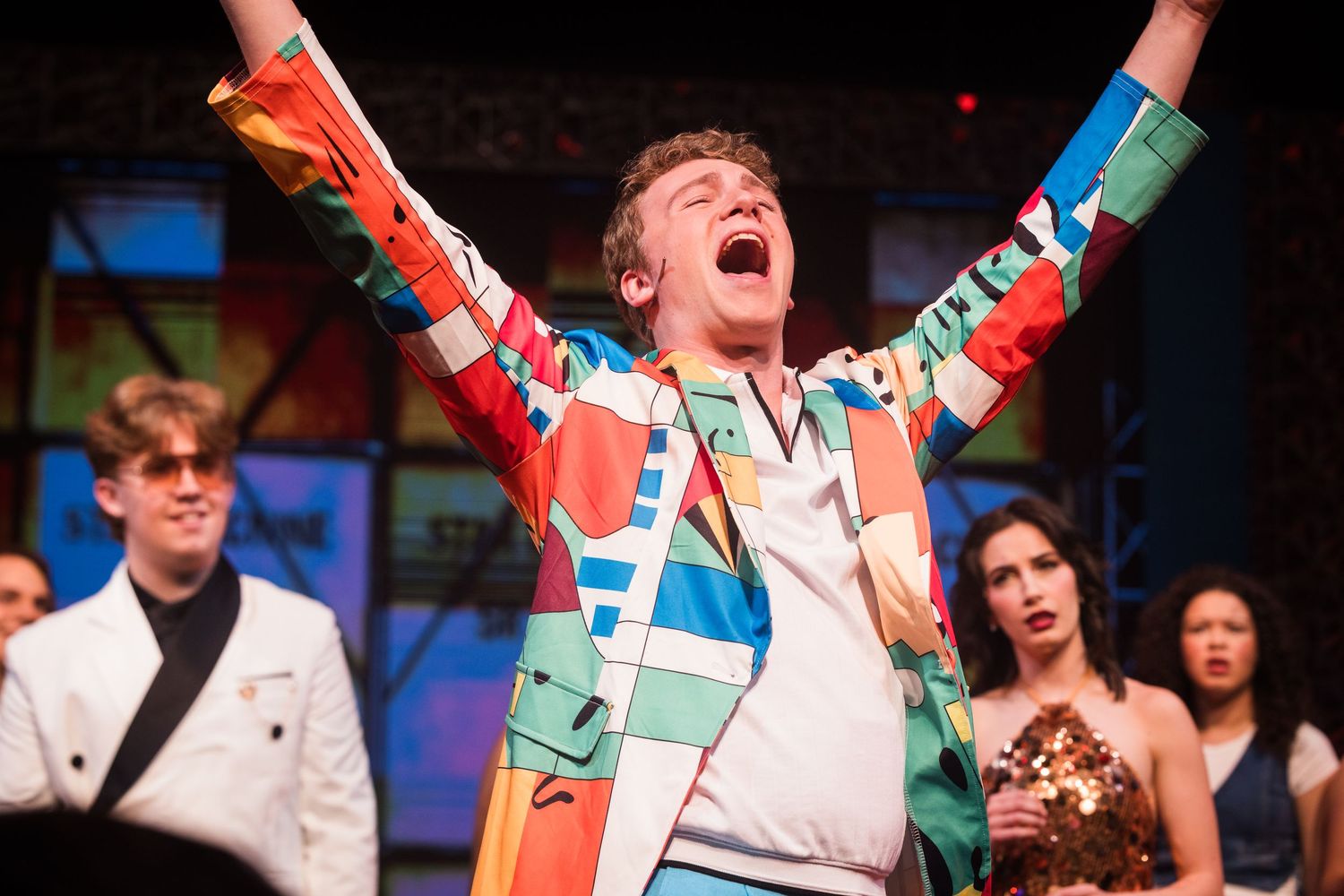 Exuberant male performer in colorful geometric blazer celebrates with arms raised on theatrical stage.