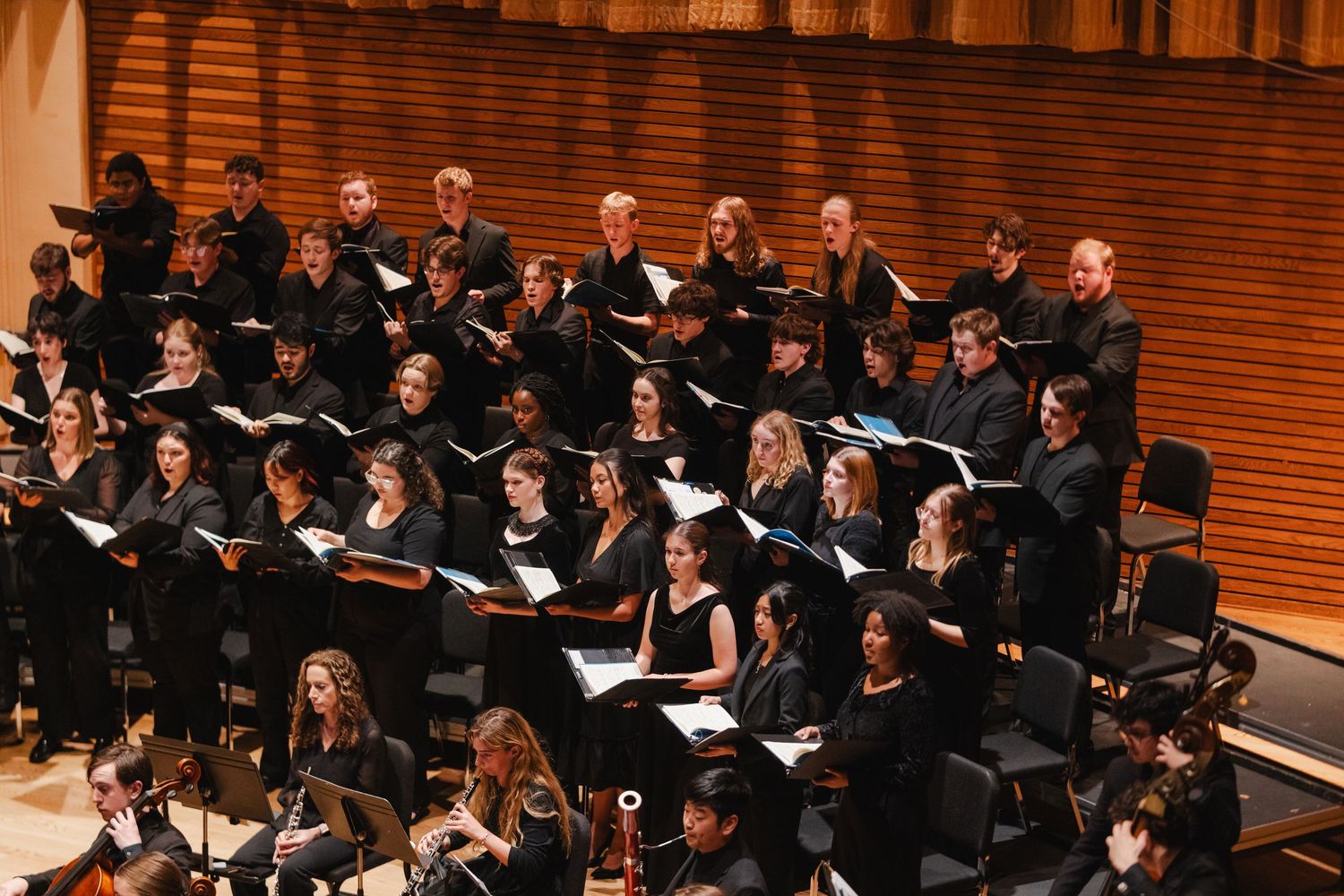 Large university choir in black robes performs with orchestra in a professional concert hall setting.