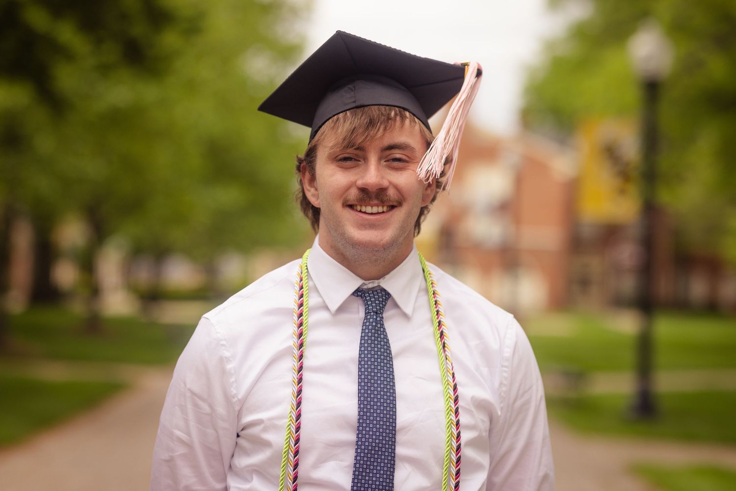 Smiling young man with mustache wearing graduation cap and colorful honor cords outdoors.