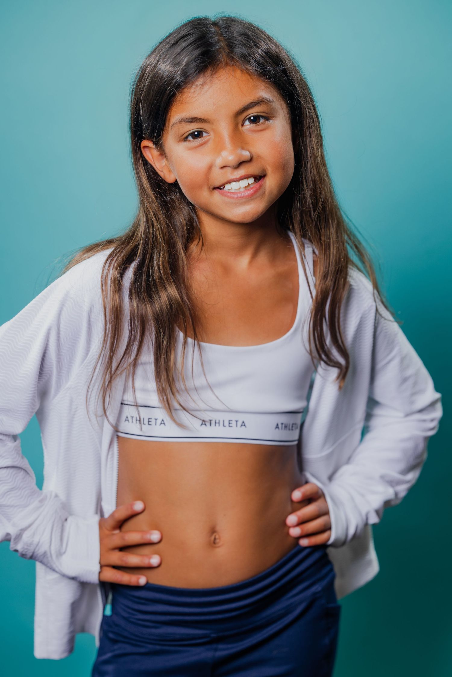 Young girl in Athleta sports bra and white jacket posing confidently against a teal background.