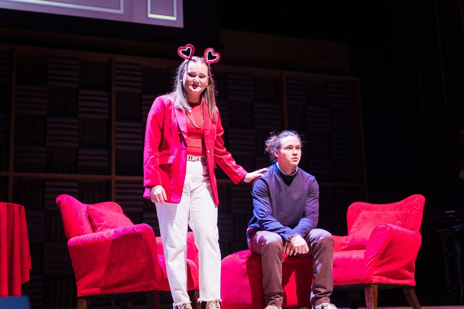 Two student actors perform on stage with red furniture during a theatrical production under stage lights.