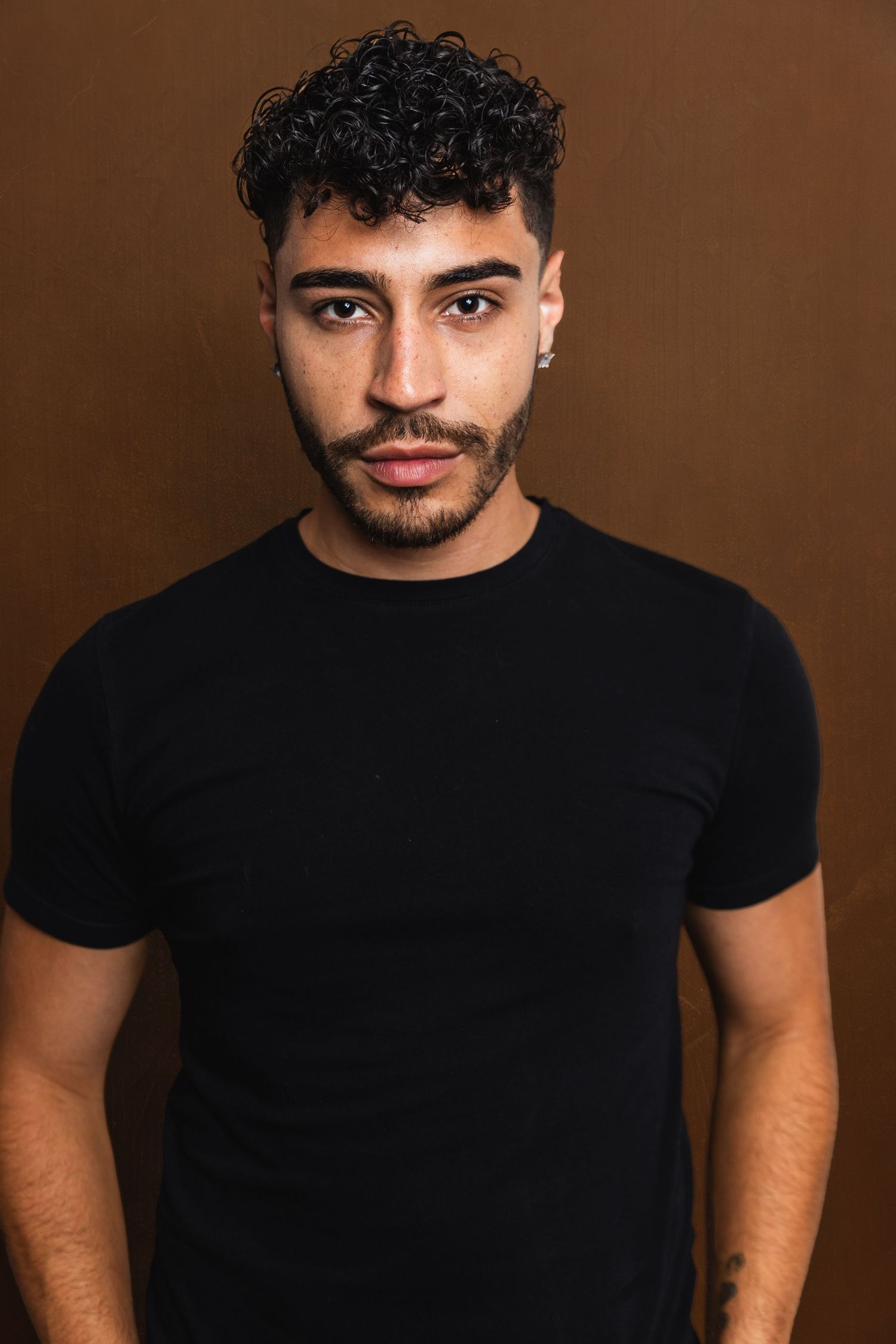 Young man with curly hair and beard in black t-shirt against brown studio background.
