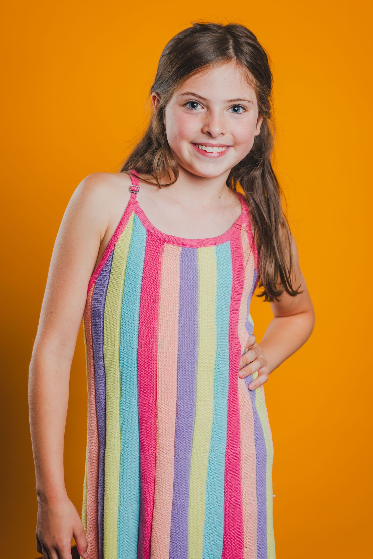 Smiling young girl in colorful rainbow striped sundress posing with hand on hip against bright orange background.