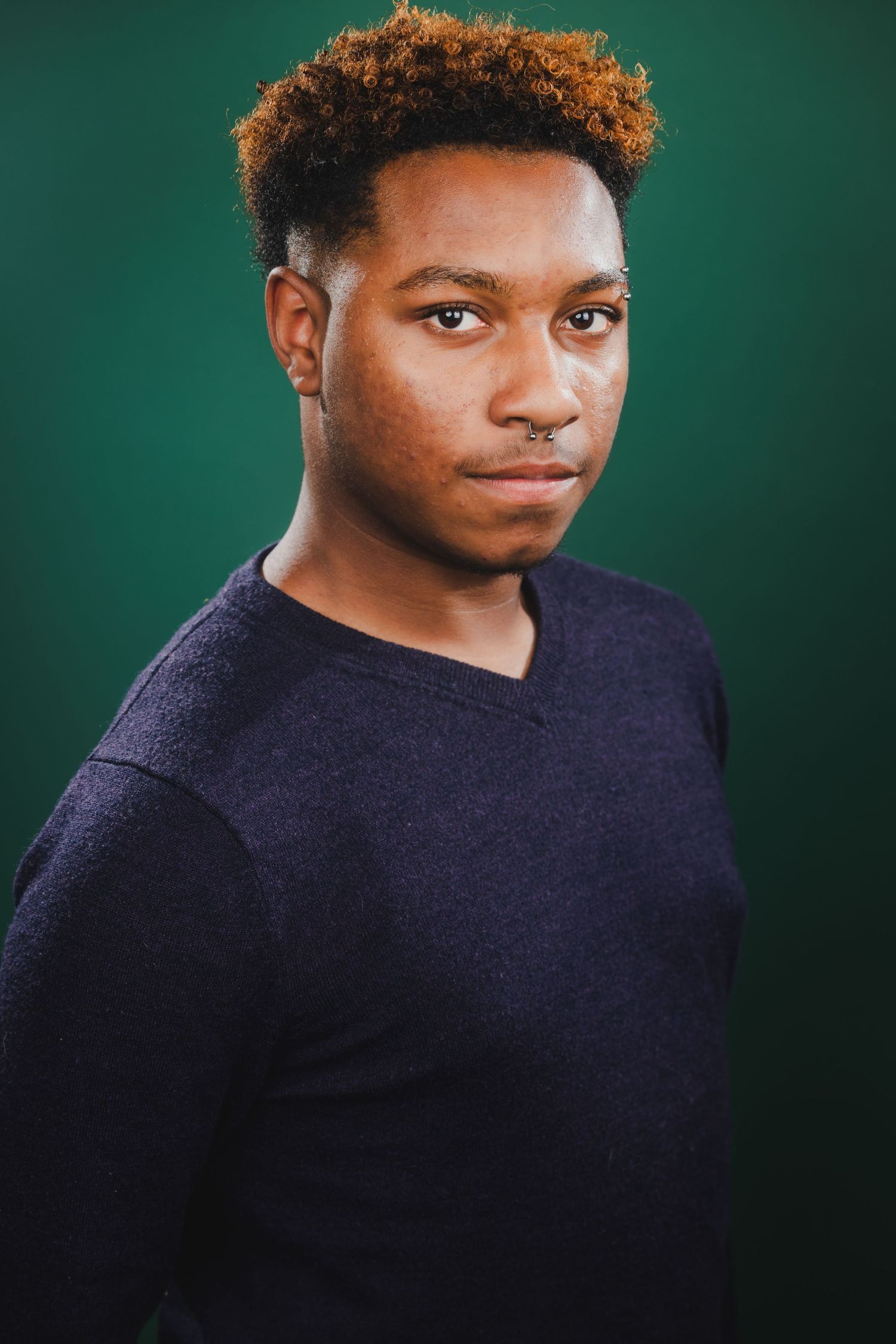 Young Black man with dyed curly hair and nose ring in a navy sweater posing against a green background.