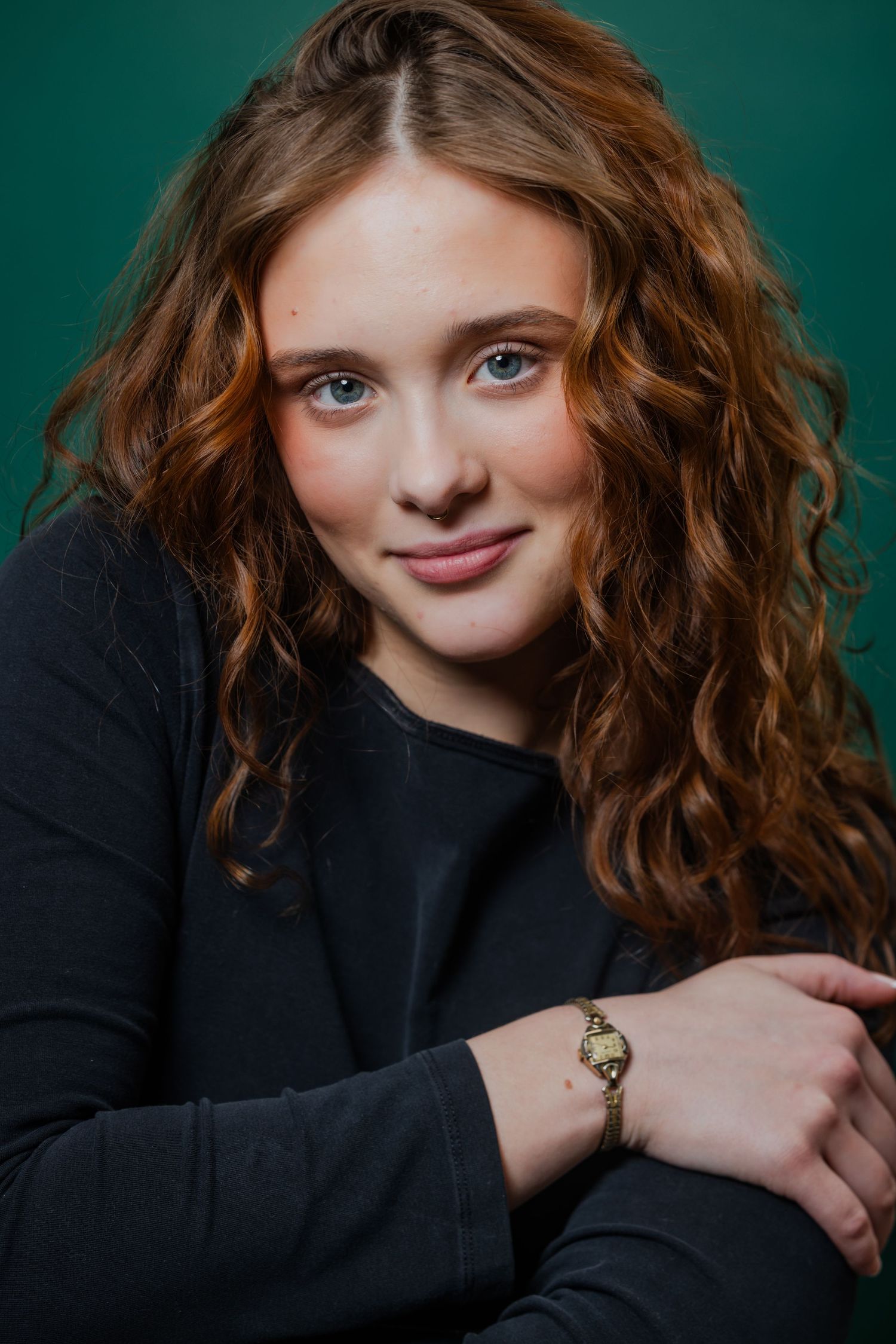 A young woman with curly auburn hair wearing a black top and gold watch poses against a green background.