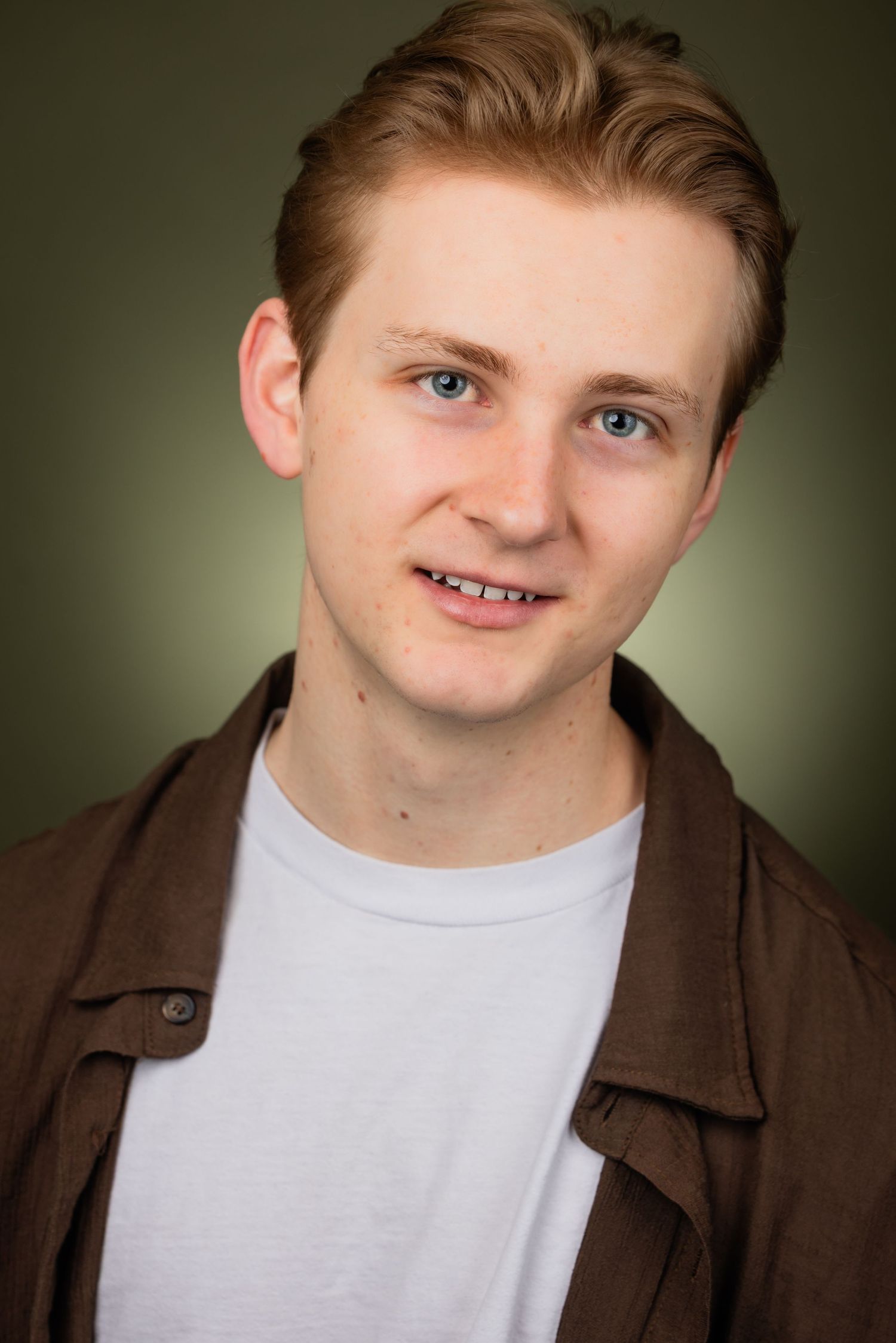 Young man with light brown hair wearing a brown jacket over white tee poses for a headshot on olive background.