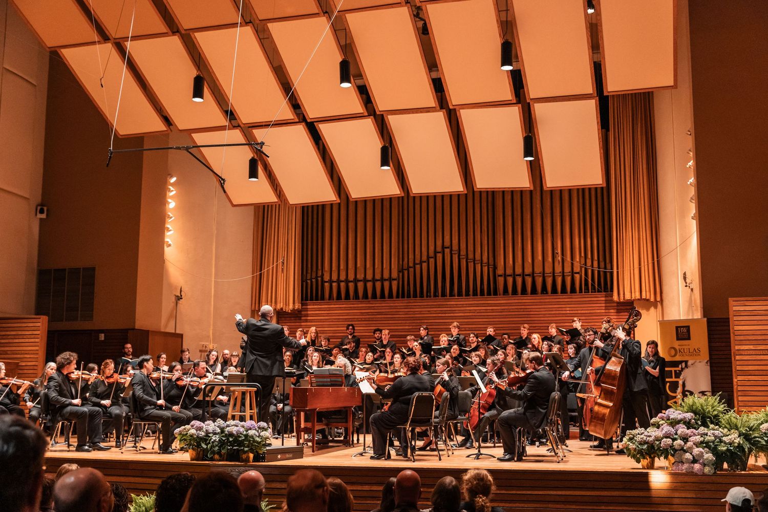 A conductor leads a full orchestra and choir ensemble in a warmly lit concert hall performance.
