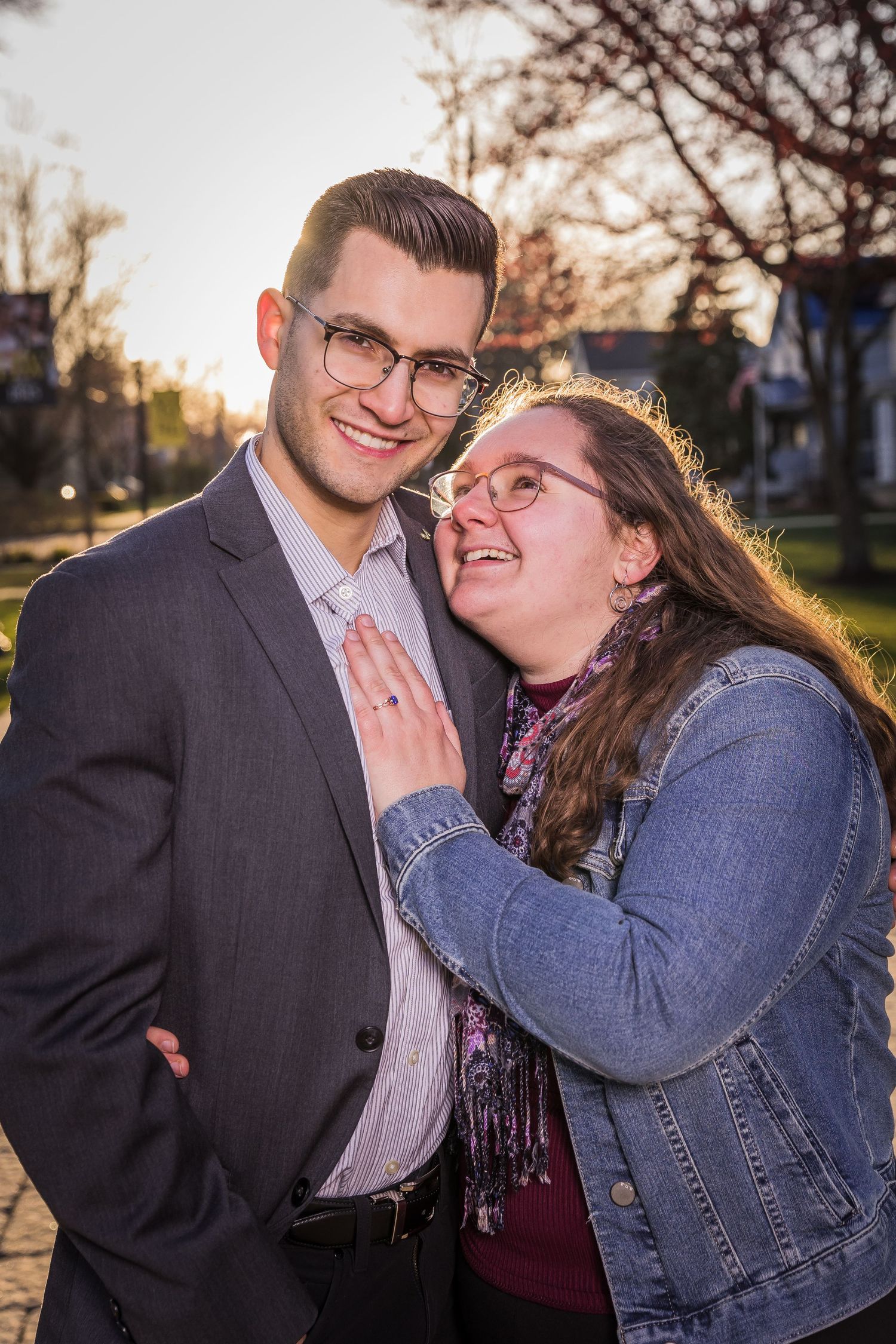 Smiling couple posing outdoors at sunset, man in suit and woman in denim jacket with glasses.