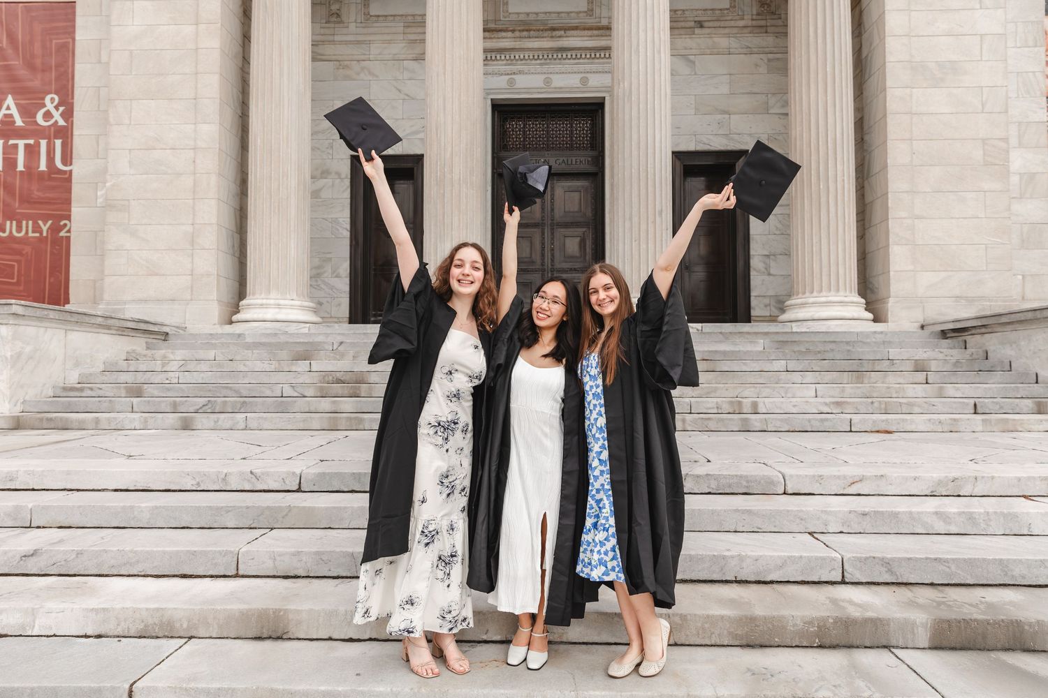 Three happy female graduates in black gowns raising caps in front of a historic building.