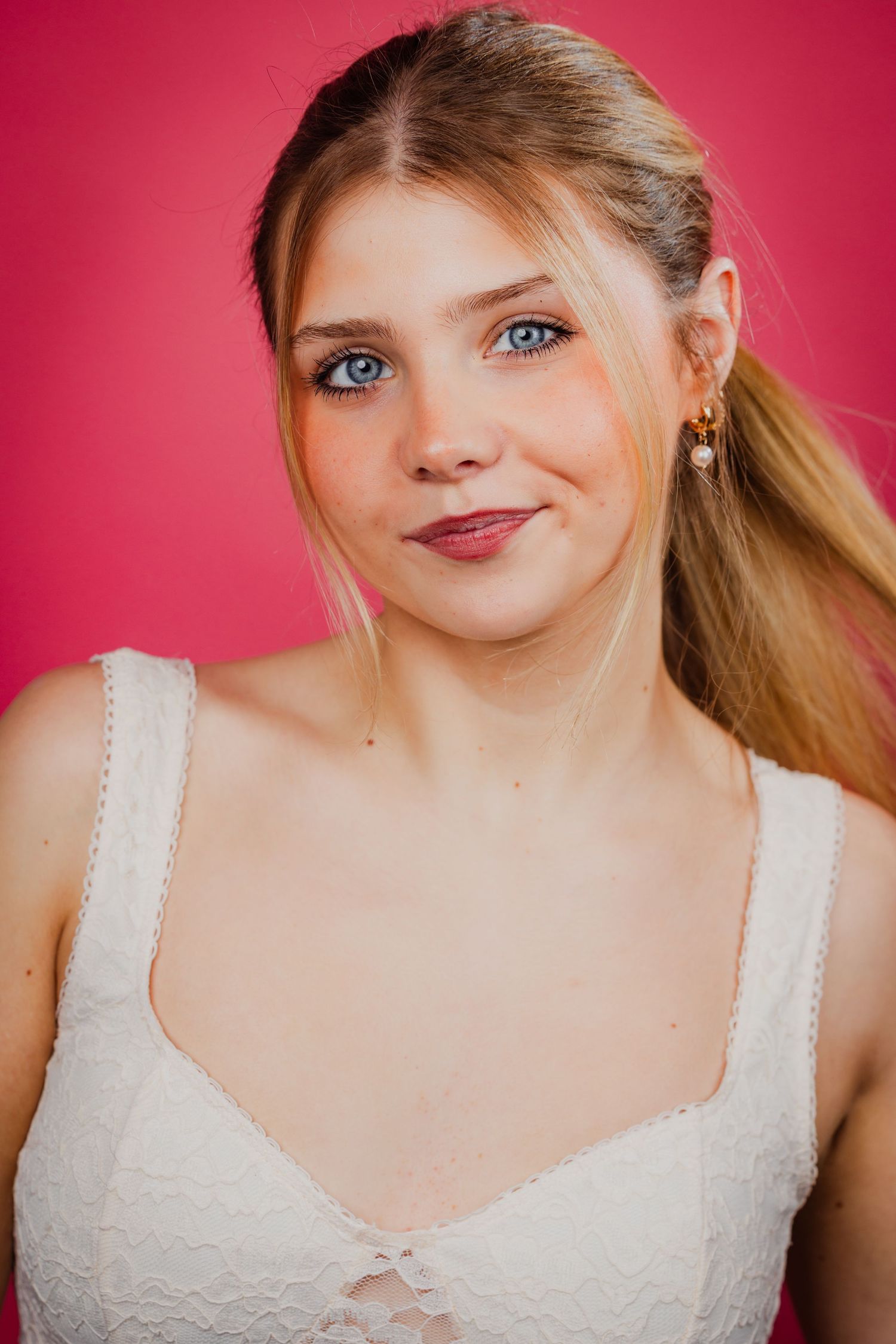 Young blonde woman with blue eyes wearing a white top and gold earrings on a pink background.