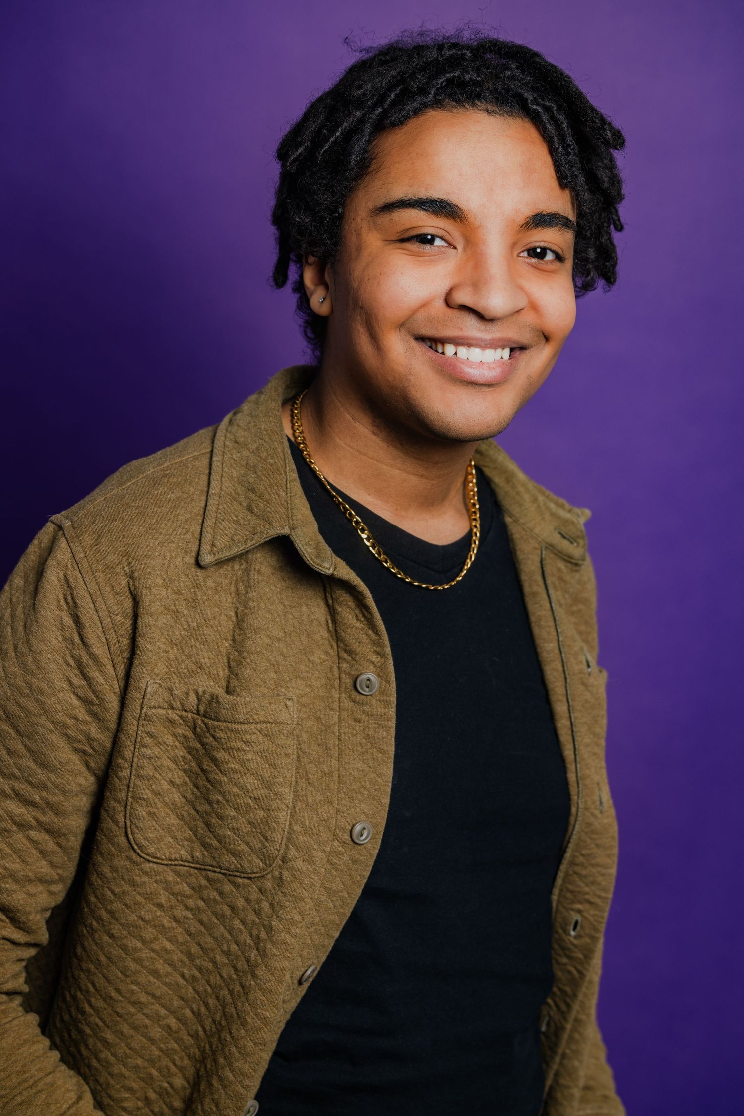Smiling young man with dreadlocks wearing olive jacket and gold chain, purple background portrait.