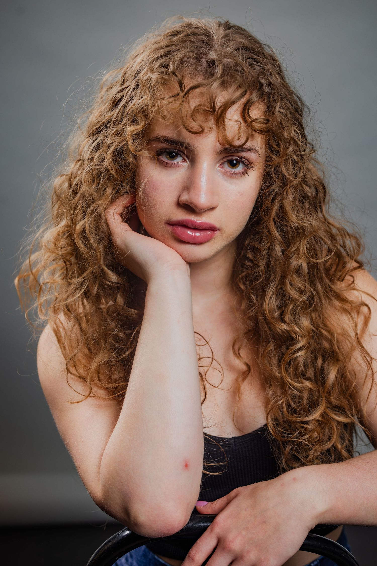 Young woman with long curly blonde hair resting her chin on her hand against a gray background.