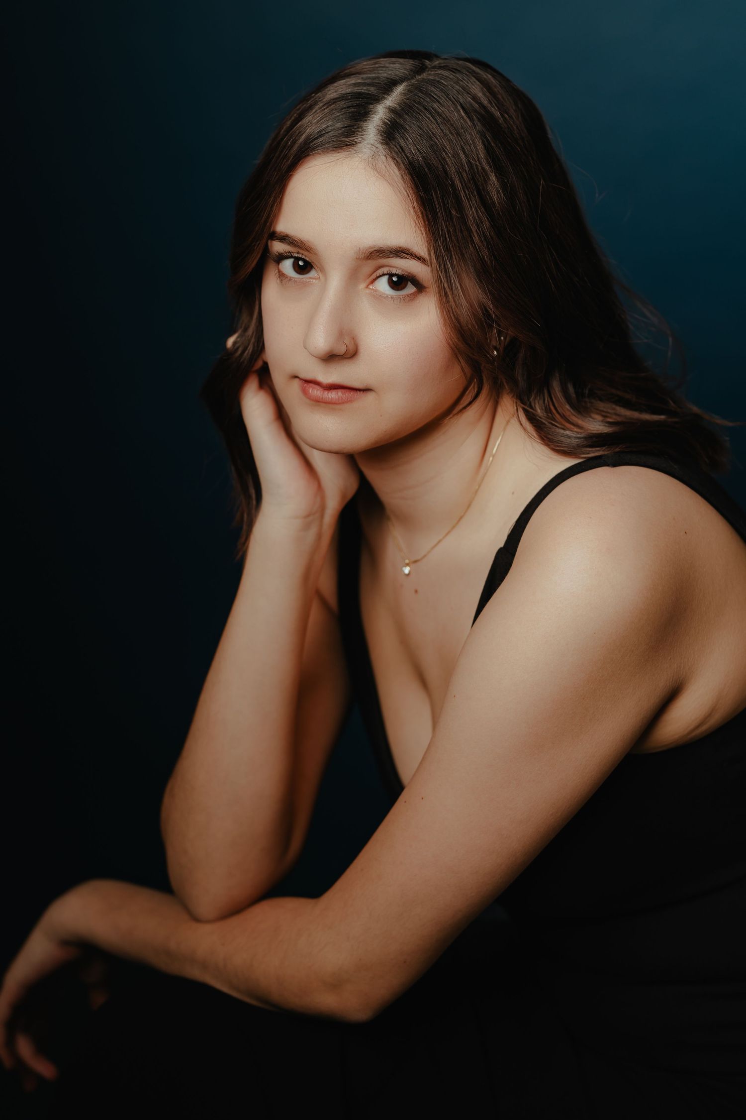 Young woman with dark hair in black top resting chin on hand against dark teal background.