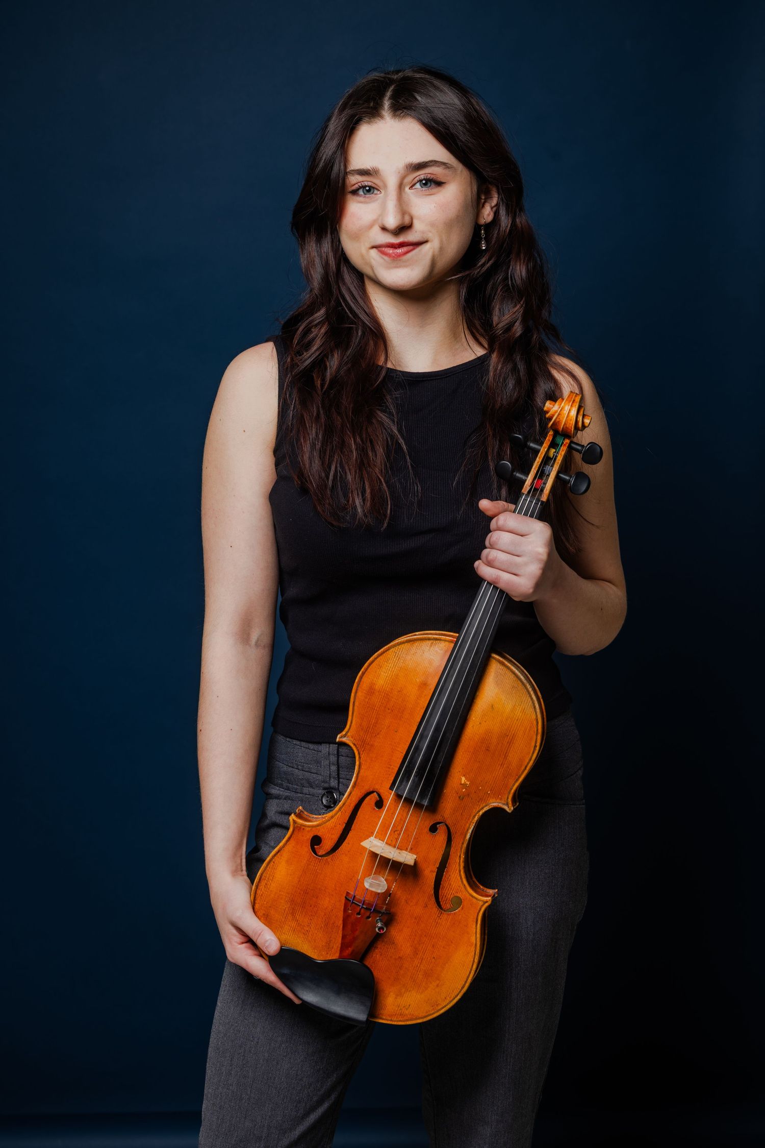 Smiling young woman in black sleeveless top holding violin against dark blue background.