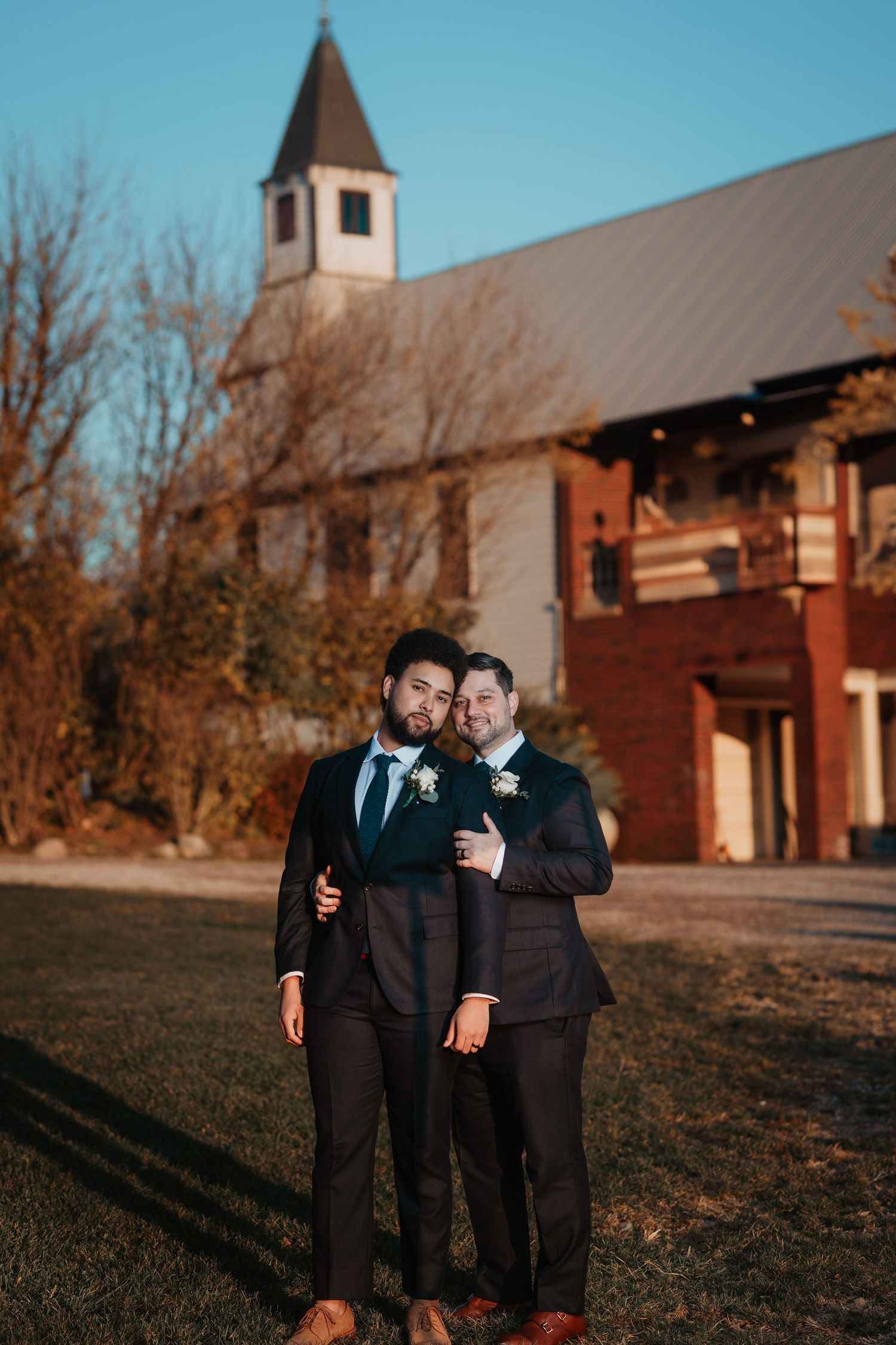 Two grooms in dark suits with boutonnieres embrace outside a rustic church venue.