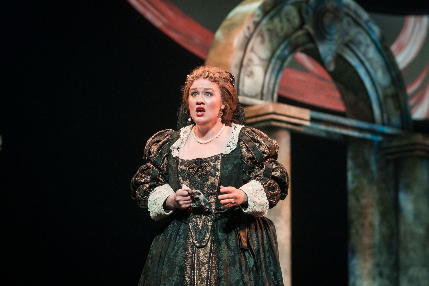 Female opera singer in an ornate dark green Renaissance gown performing passionately on a dramatic stage.