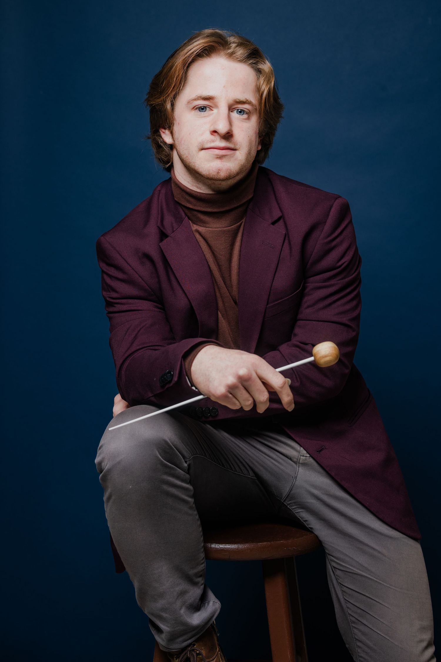 Young male conductor holding a baton while seated against a dark blue background.