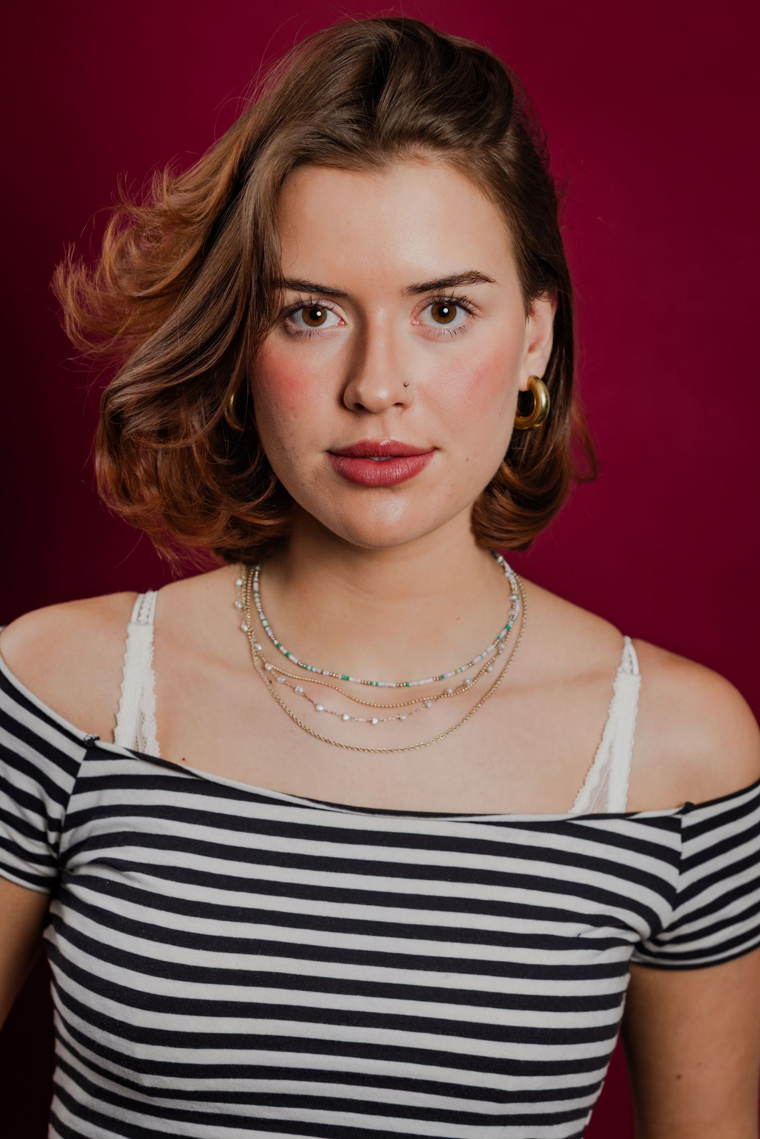 Young woman with short auburn hair wearing a striped off-shoulder top and layered necklaces on red background.