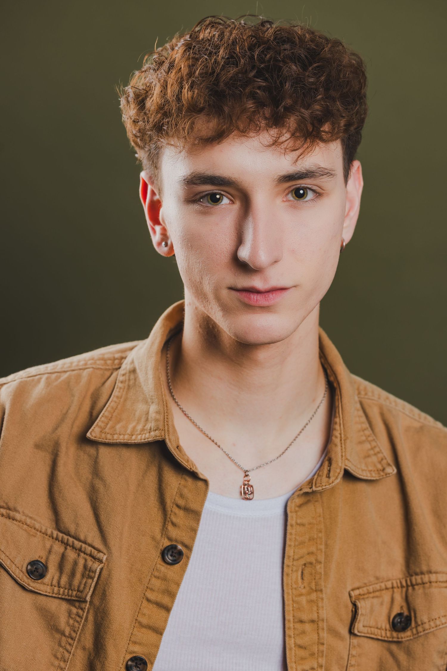 Young man with curly auburn hair wearing tan jacket and gold pendant necklace on olive green background.