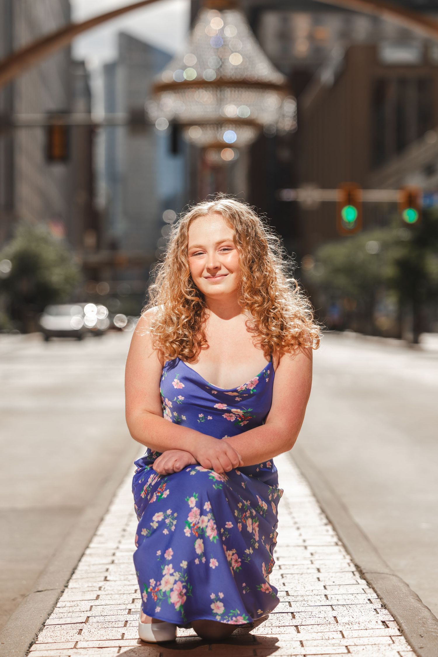 Young woman with curly blonde hair in blue floral dress kneeling on a sunlit city street.