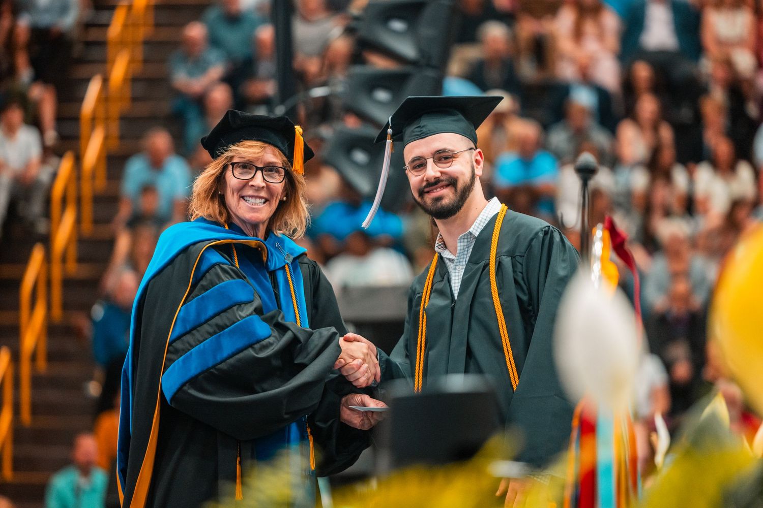 Graduate in cap and gown shaking hands with a faculty member in blue academic regalia at commencement.