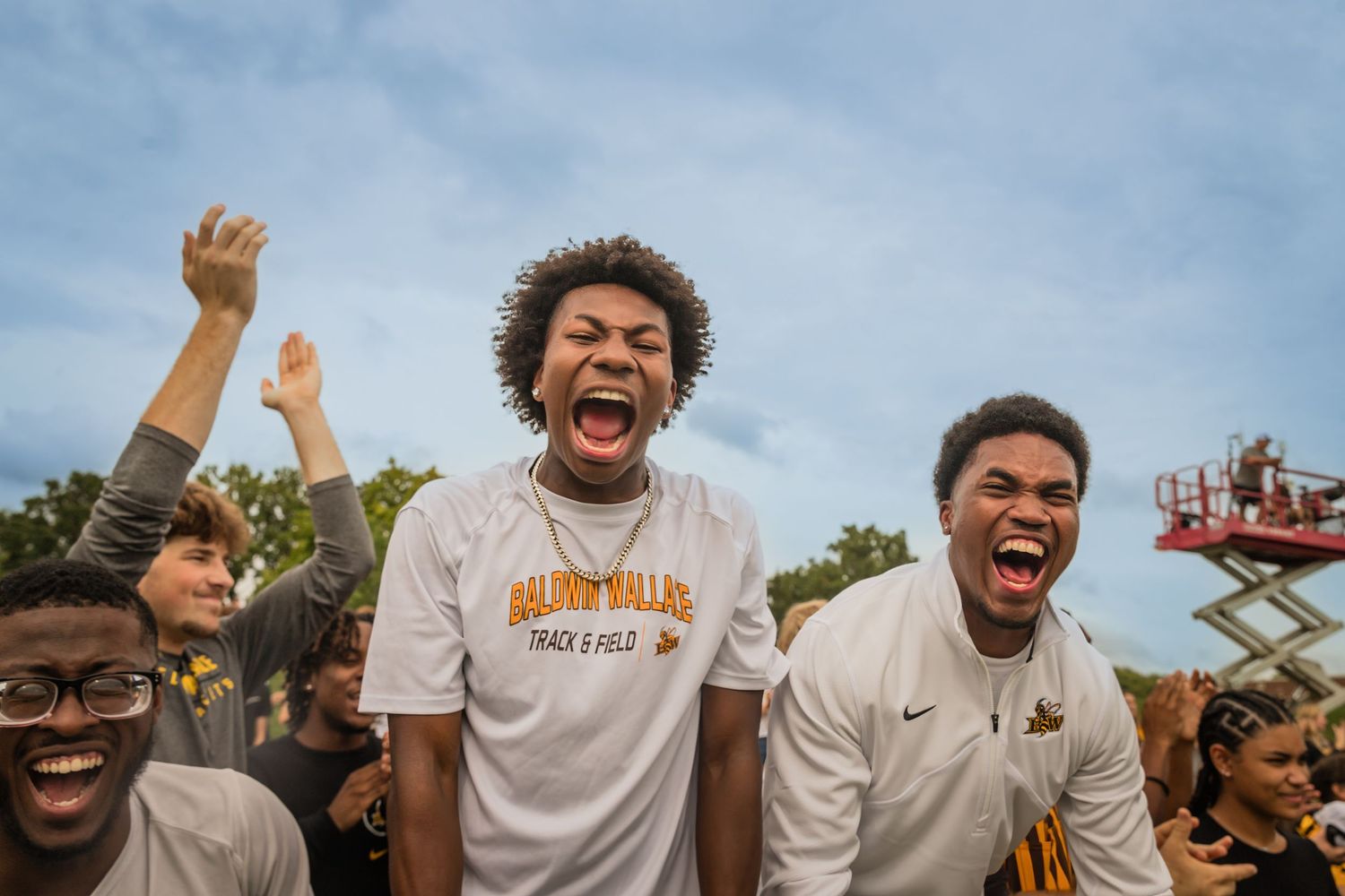 Three excited young men cheer enthusiastically outdoors at a school sporting event.