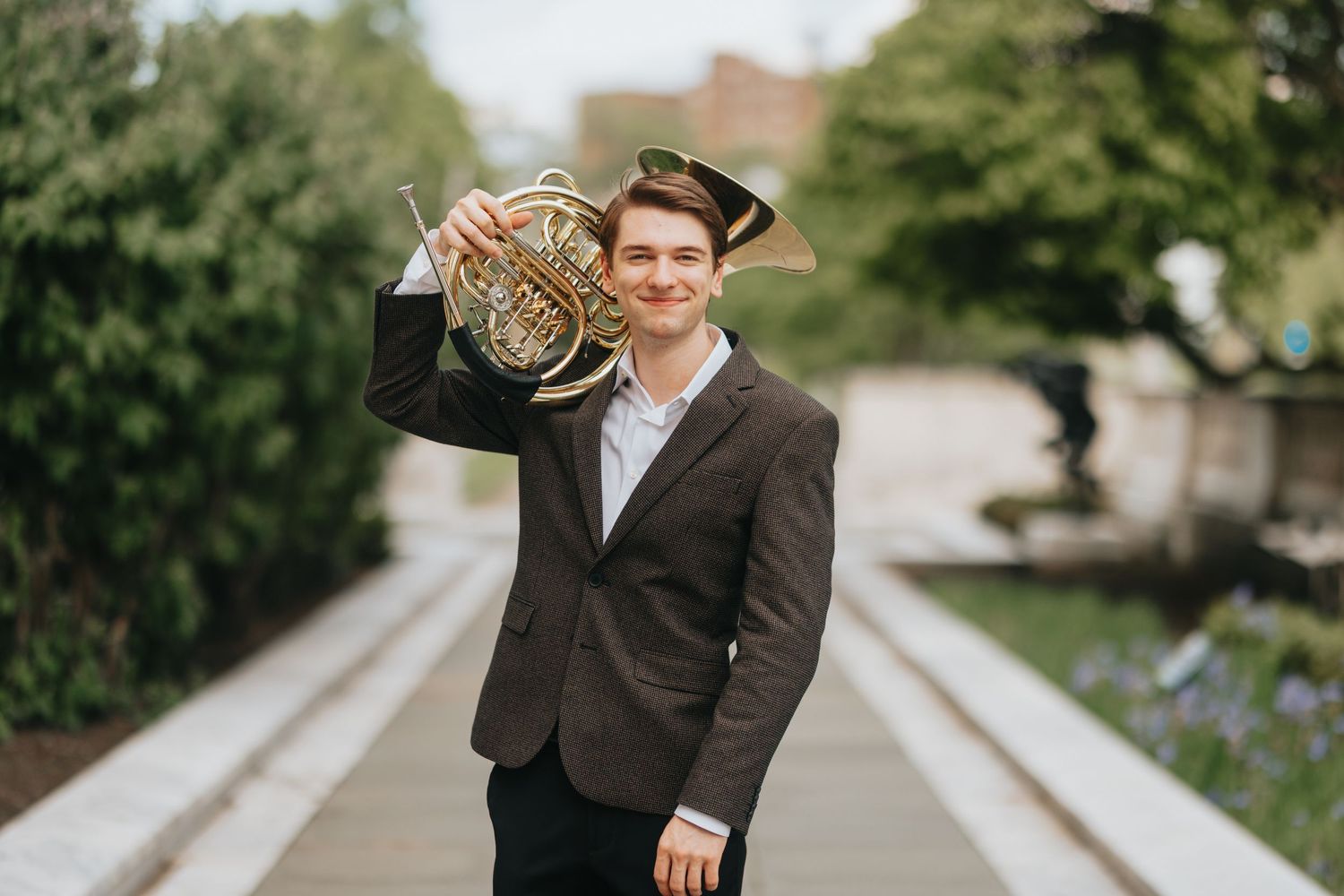 Smiling young musician holds a French horn outdoors in a lush green park setting.