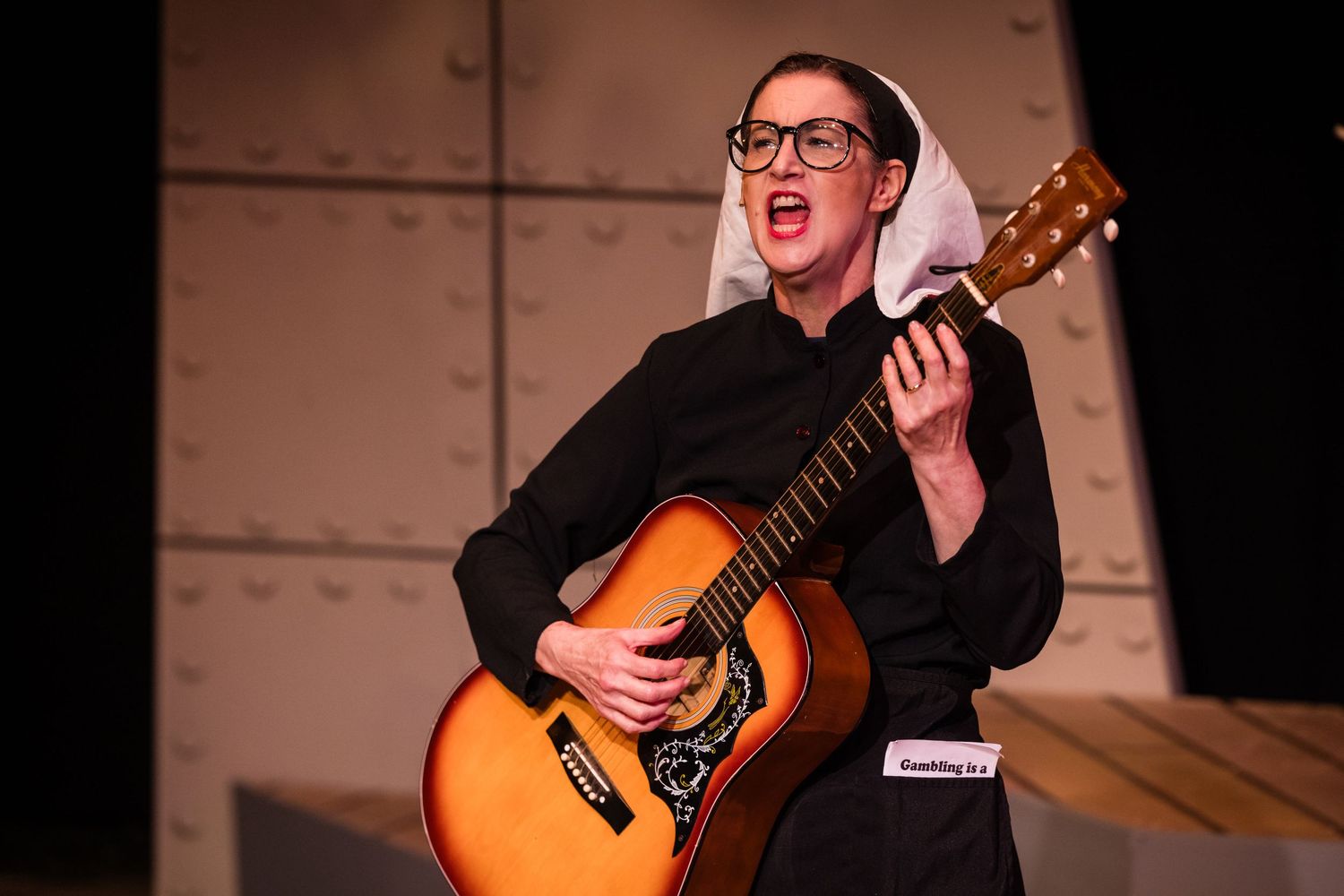 Performer dressed as a nun playing acoustic guitar and singing on a dark stage.