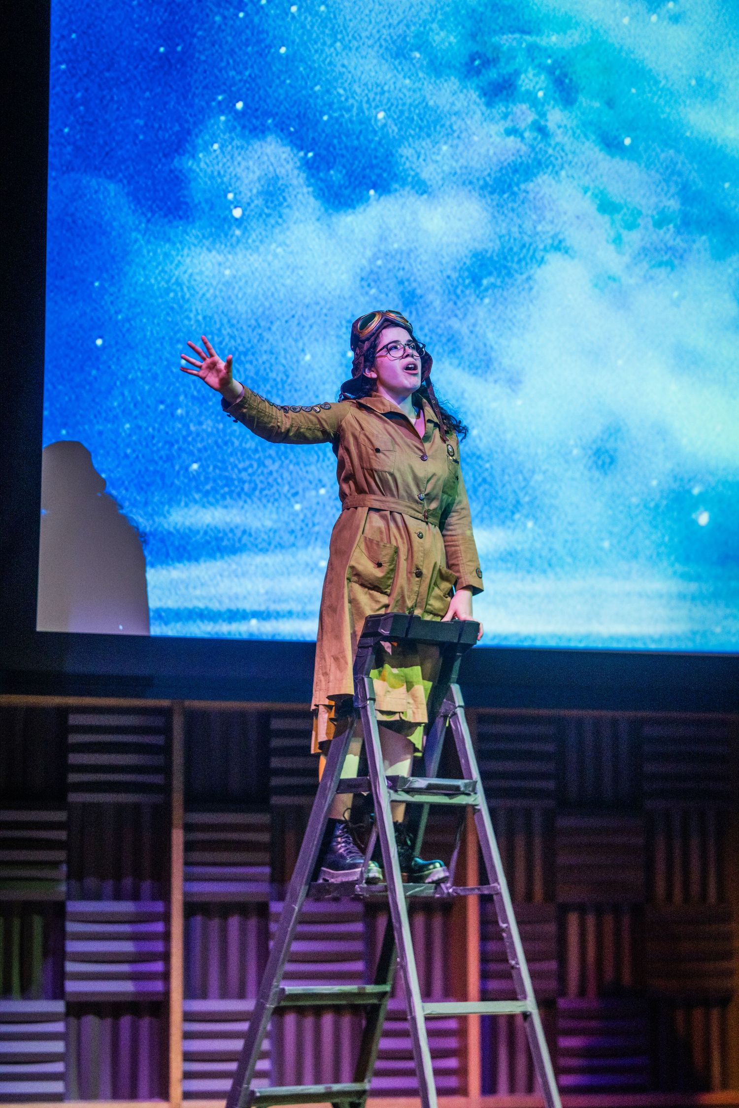 Performer in a tan trench coat stands atop a ladder reaching upward on a stage with a starry blue backdrop.