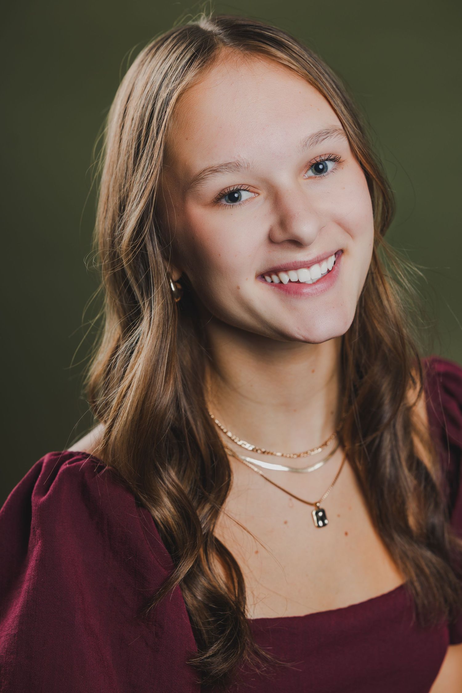 Smiling teenage girl with long blonde hair wearing a burgundy top and layered necklaces on dark background.