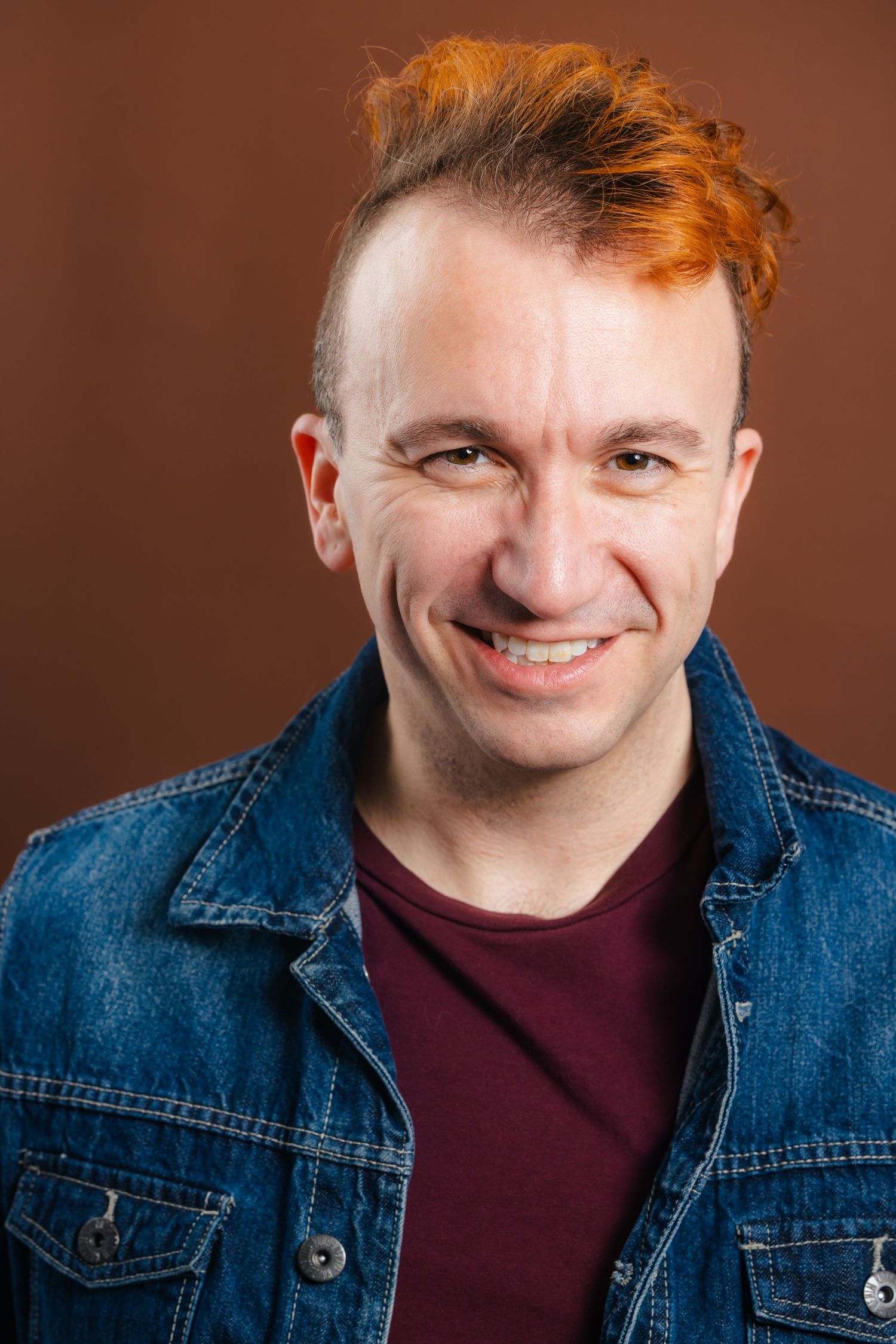 Smiling man with red mohawk hairstyle wearing denim jacket over maroon shirt, studio portrait.