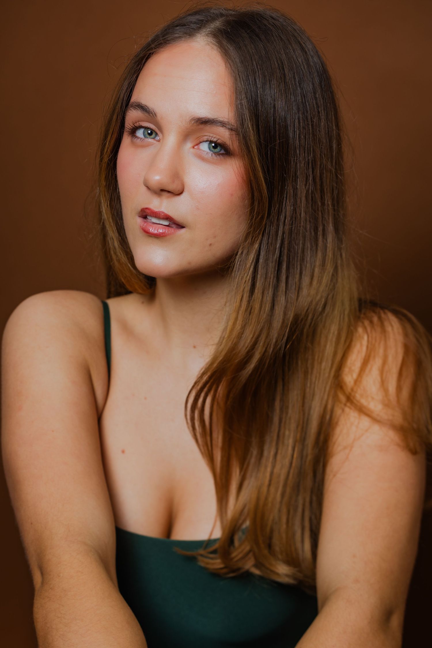 A young woman with long brown hair wearing a green spaghetti-strap top poses against a brown background.