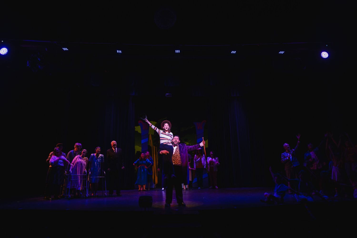 A theatrical ensemble performs on a dimly lit stage with a central performer raising one arm dramatically.