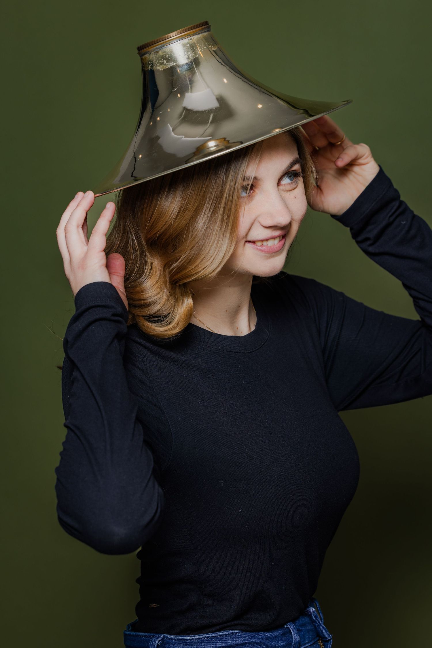 Playful woman balancing a metallic lamp shade on her head, wearing a black top on a green background.