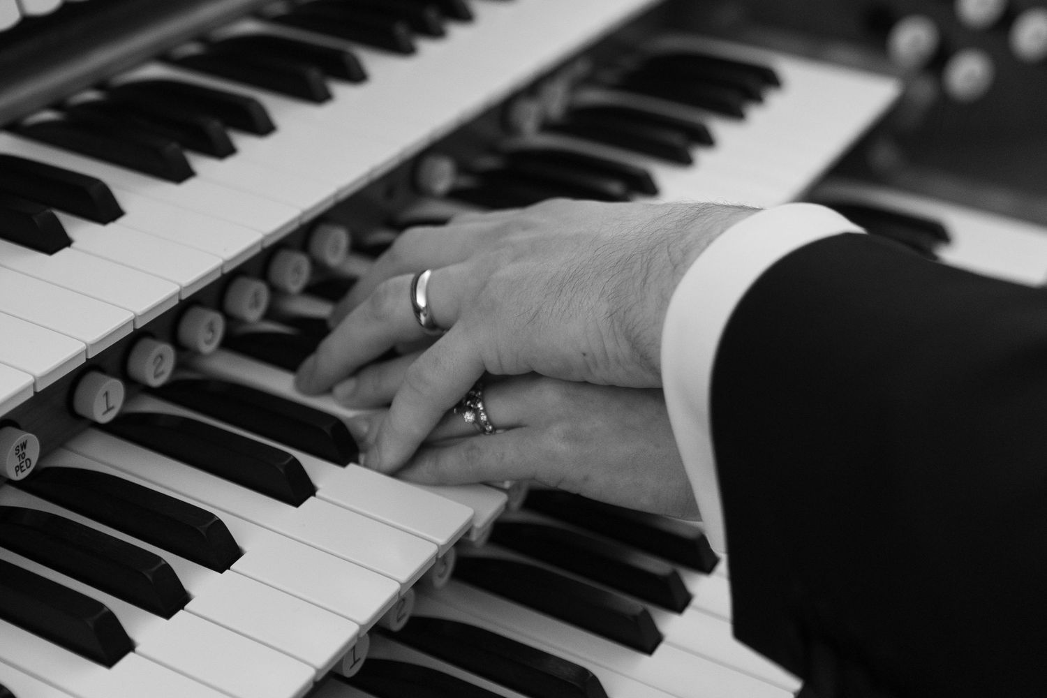 Black and white close-up of hands playing a piano organ with wedding ring visible.