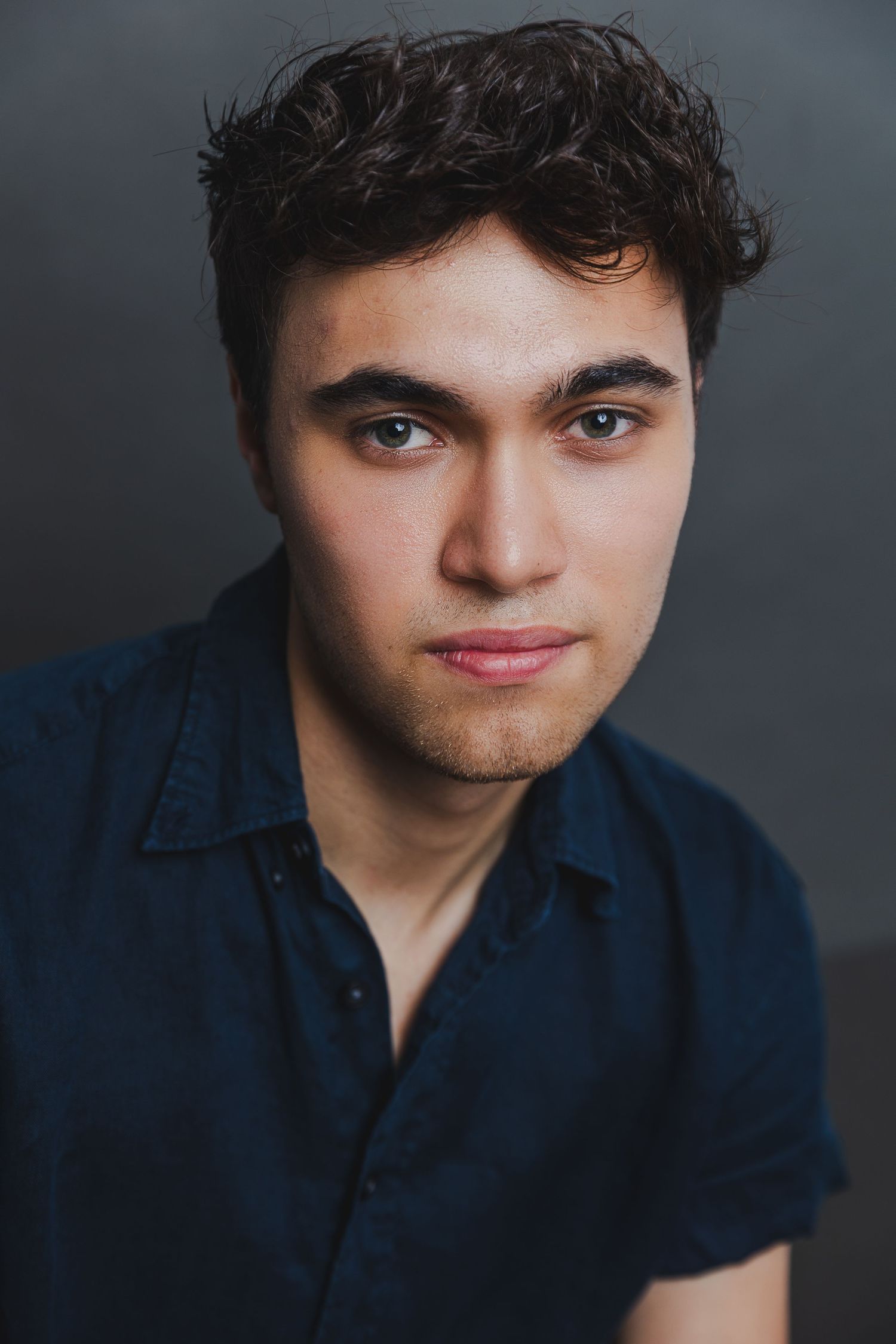 Young man with dark curly hair in navy blue shirt posing against dark gray background.