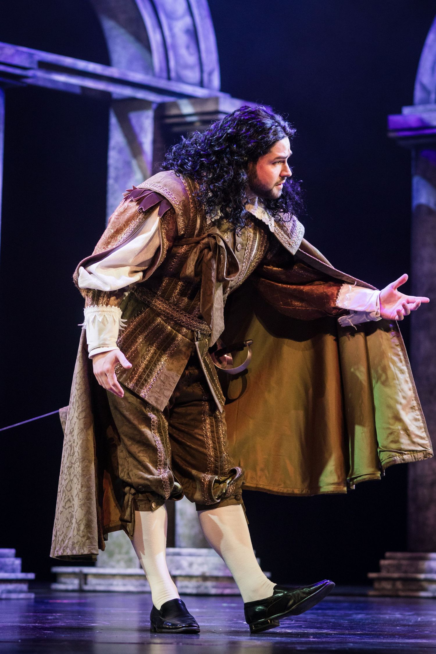 Male performer in brown period costume with long curly black hair gesturing on a dimly lit opera stage.