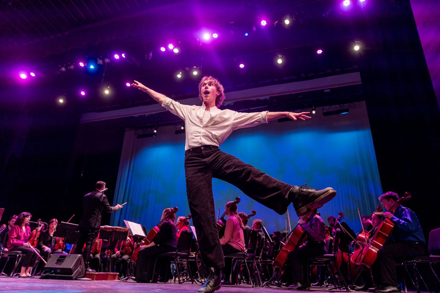 A dancer leaps dramatically in front of an orchestra under vibrant purple stage lighting.