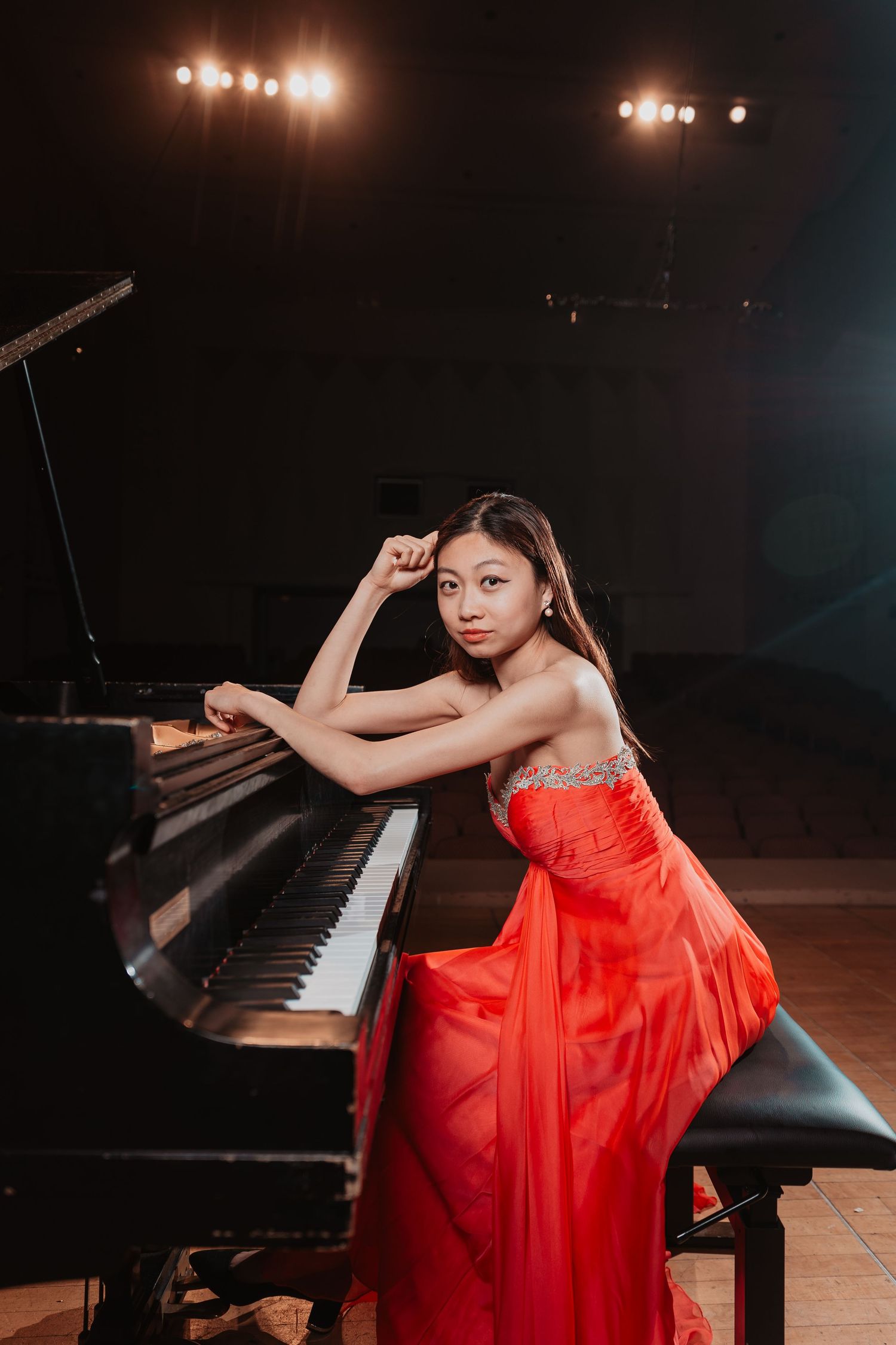 Elegant young woman in a red gown leaning on a grand piano on a concert stage under dramatic spotlights.