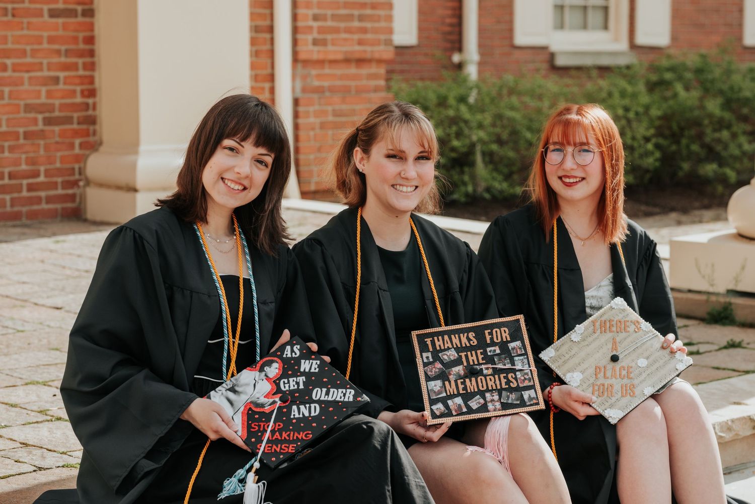 Three female graduates in black gowns sitting together holding decorated graduation caps outdoors.