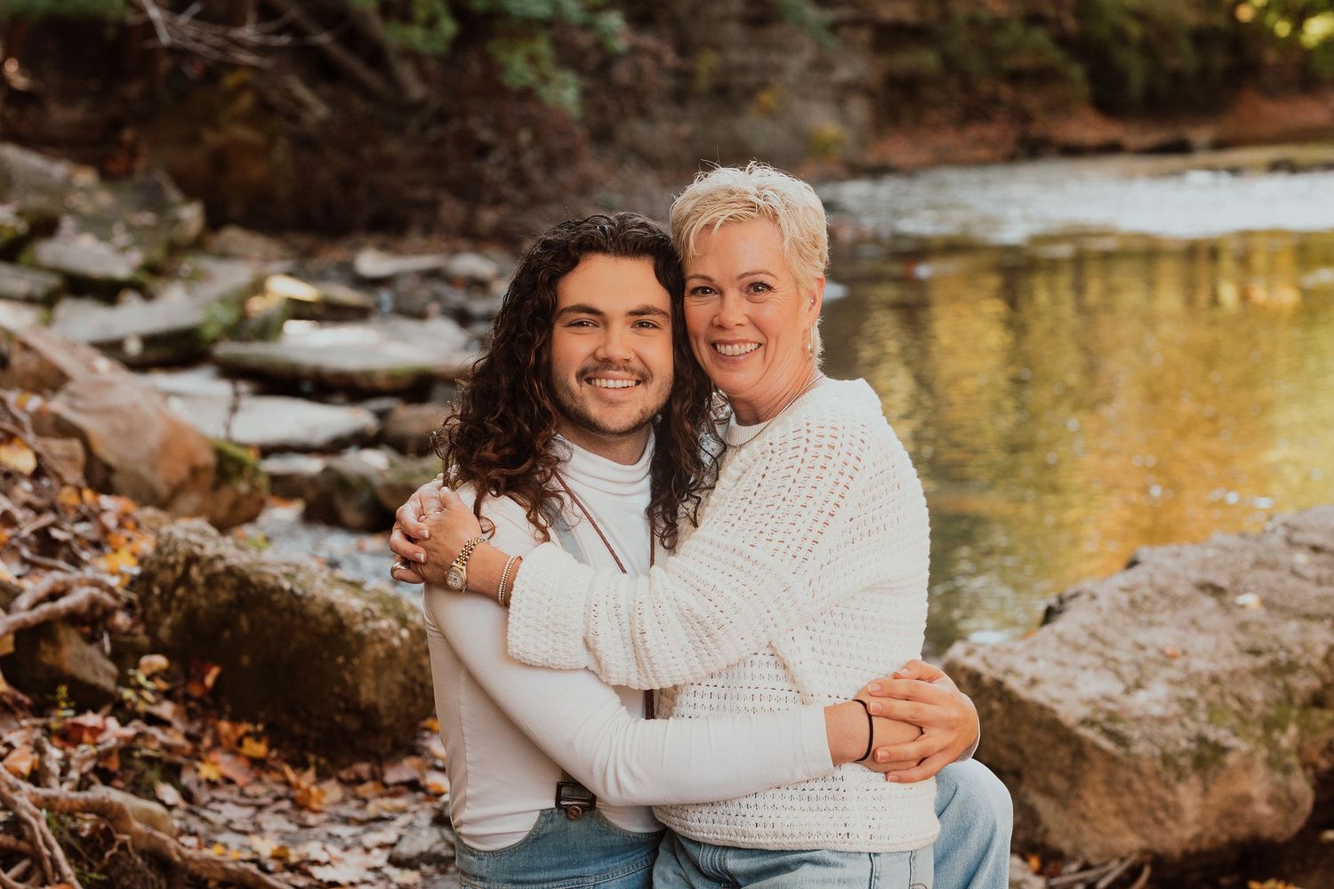 Mother and son embracing warmly beside a rocky creek in autumn outdoor setting.