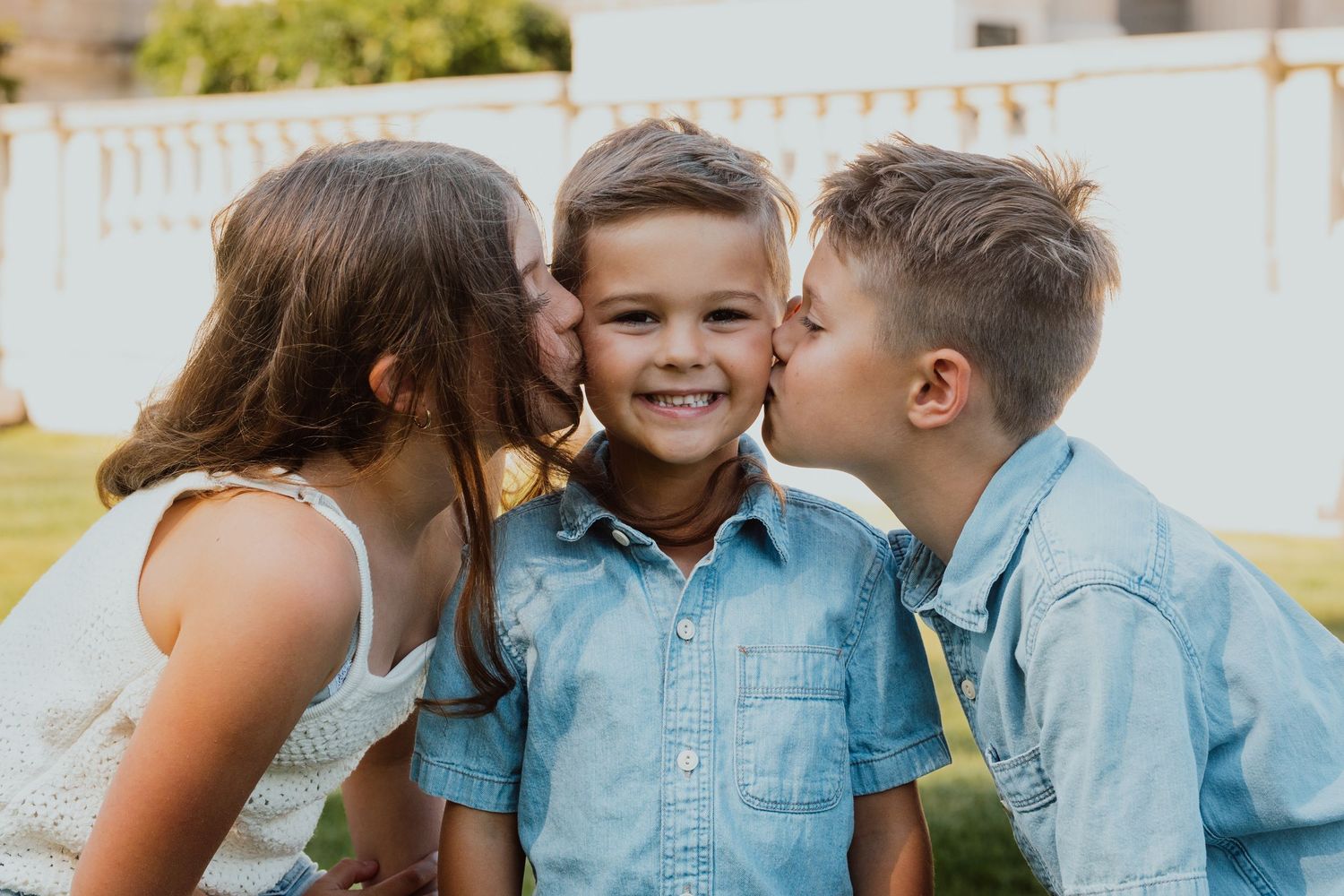 Three young siblings share sweet cheek kisses outdoors in a heartwarming portrait.