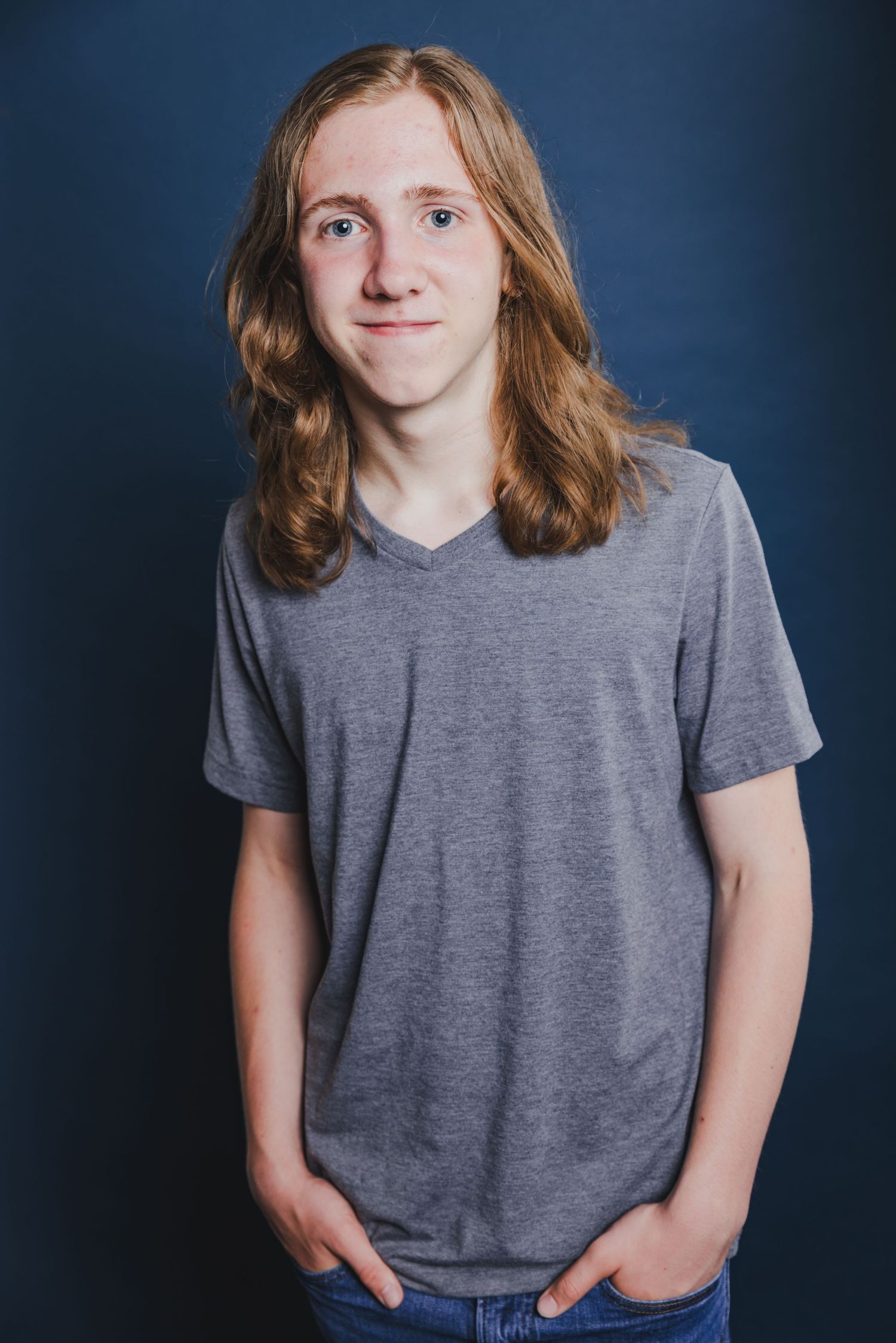 Teenage boy with long auburn wavy hair in a gray t-shirt posing against a dark blue background.