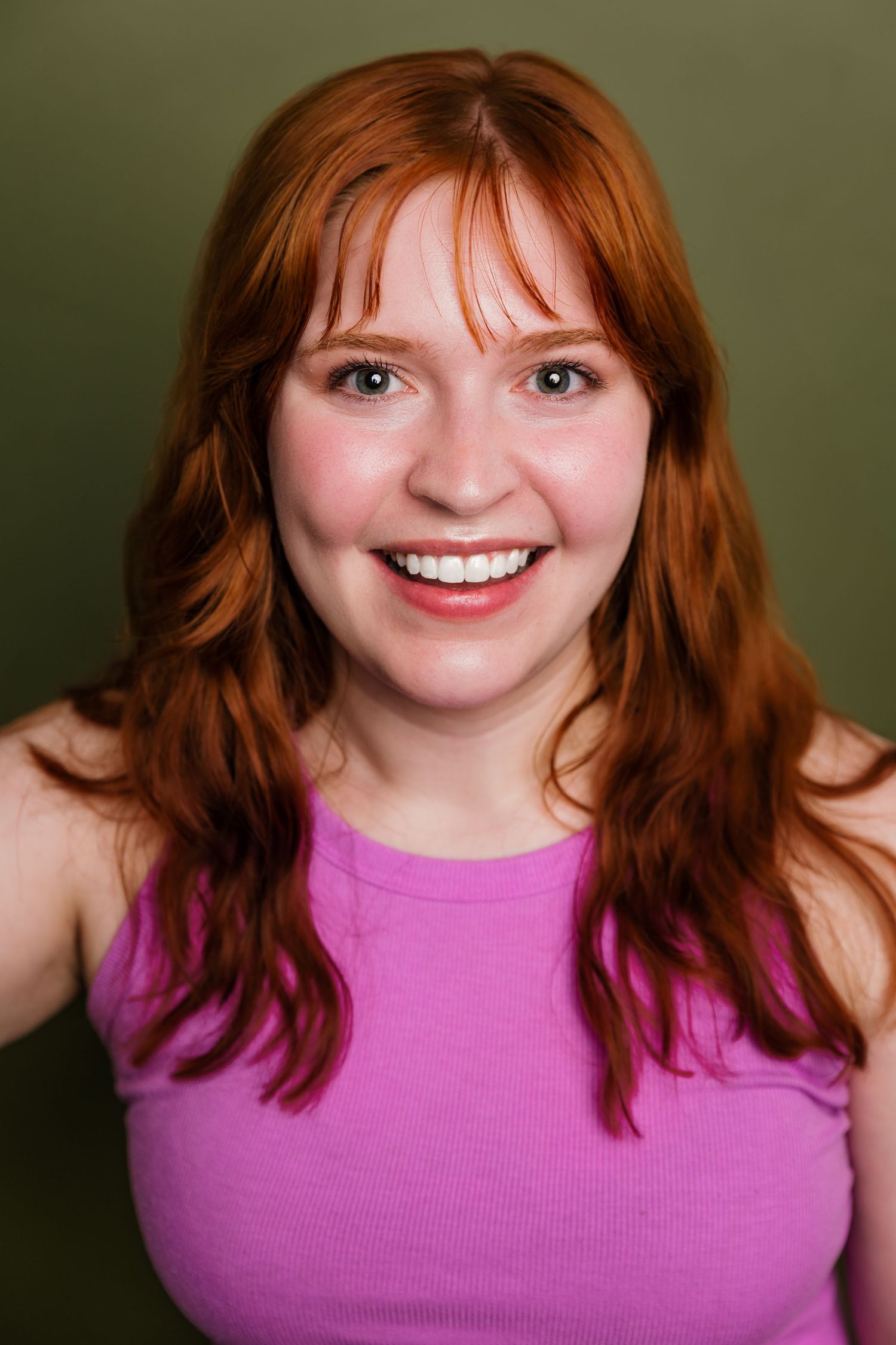 Smiling redhead woman in pink tank top posing against an olive green background.