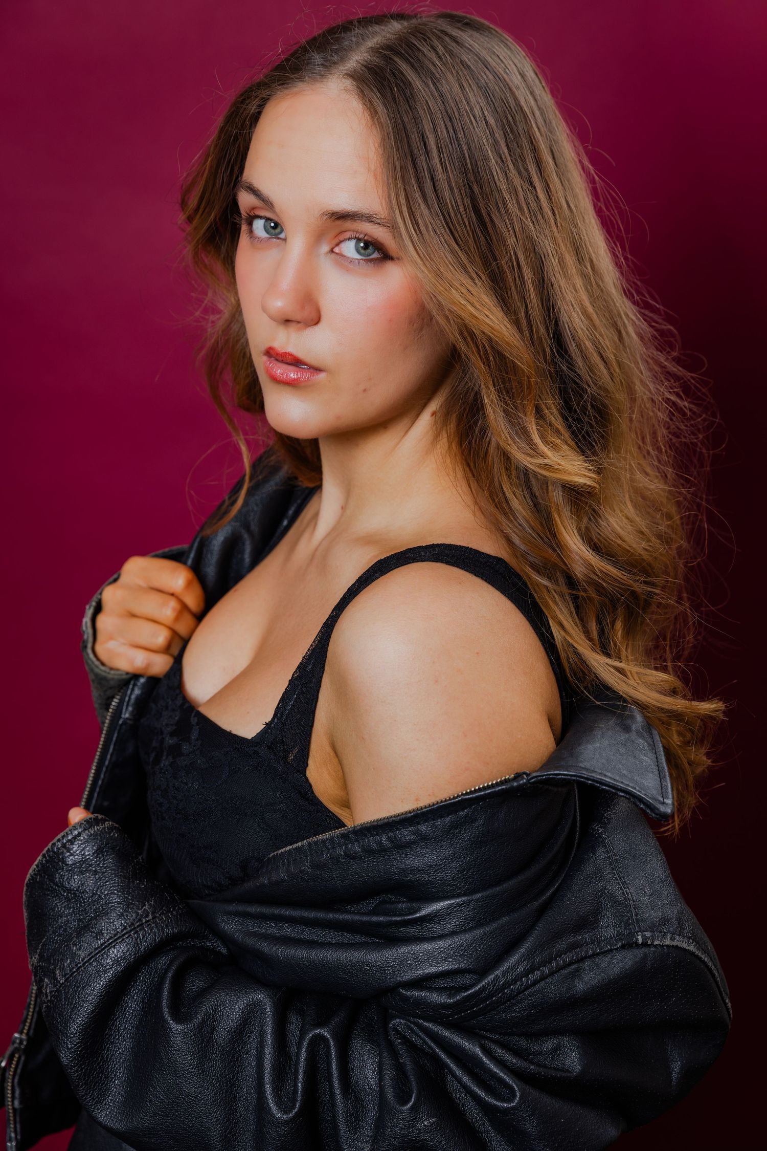 Young woman with long blonde hair posing in a black top and leather jacket against a dark red background.