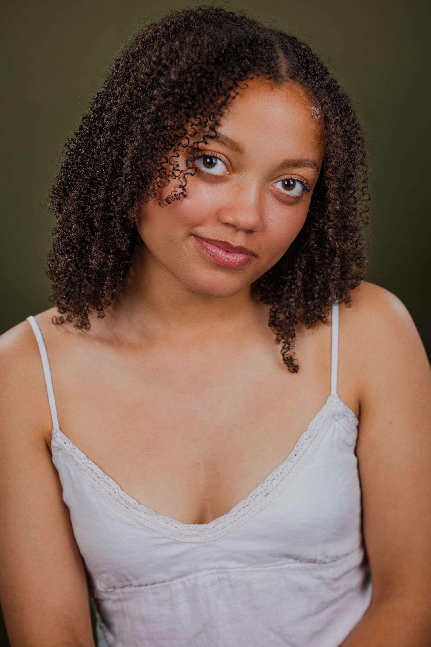 Young woman with curly brown hair wearing a white camisole, smiling softly against a green background.