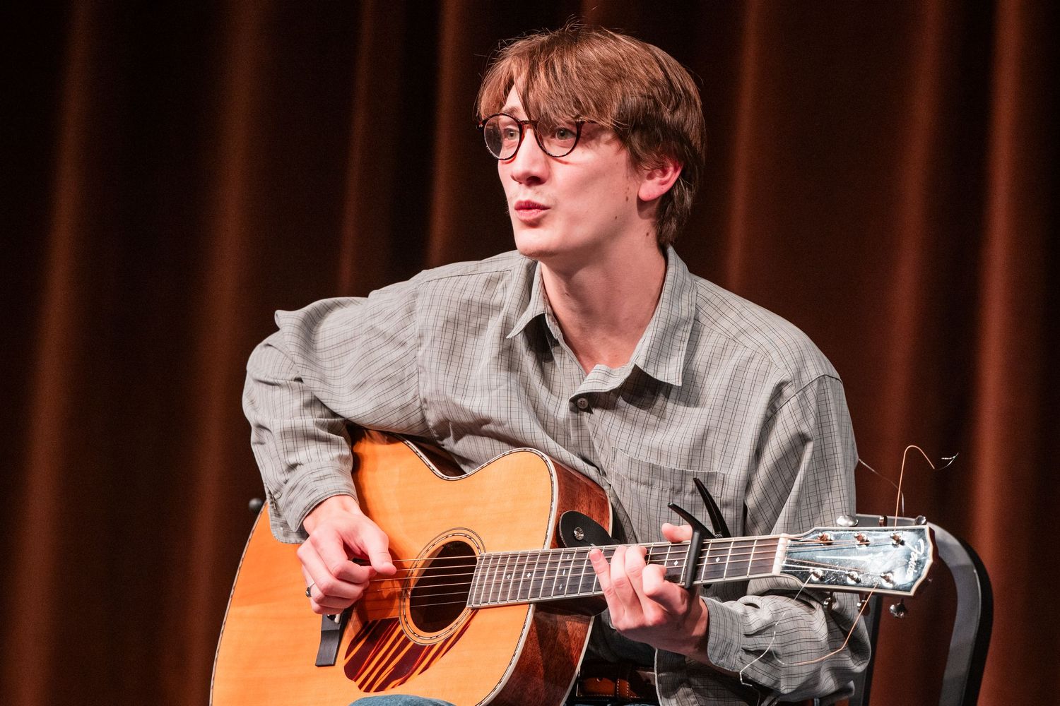 Young male musician with glasses plays acoustic guitar while seated on a theater stage.