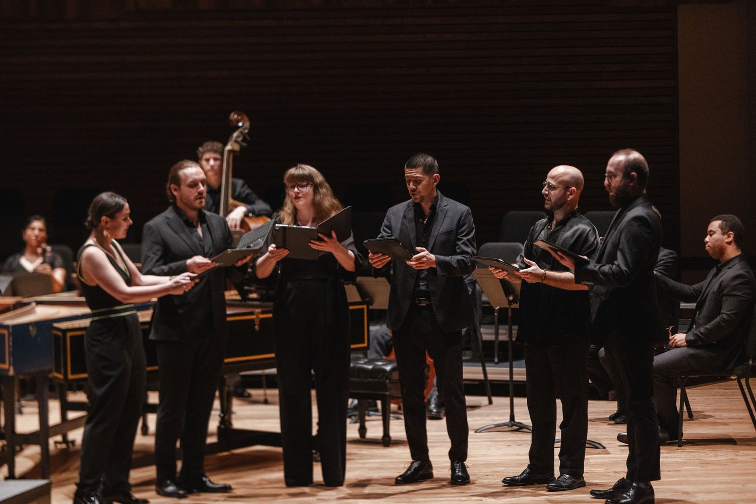 Chamber ensemble performers in black attire singing from scores on a concert hall stage with period instruments.