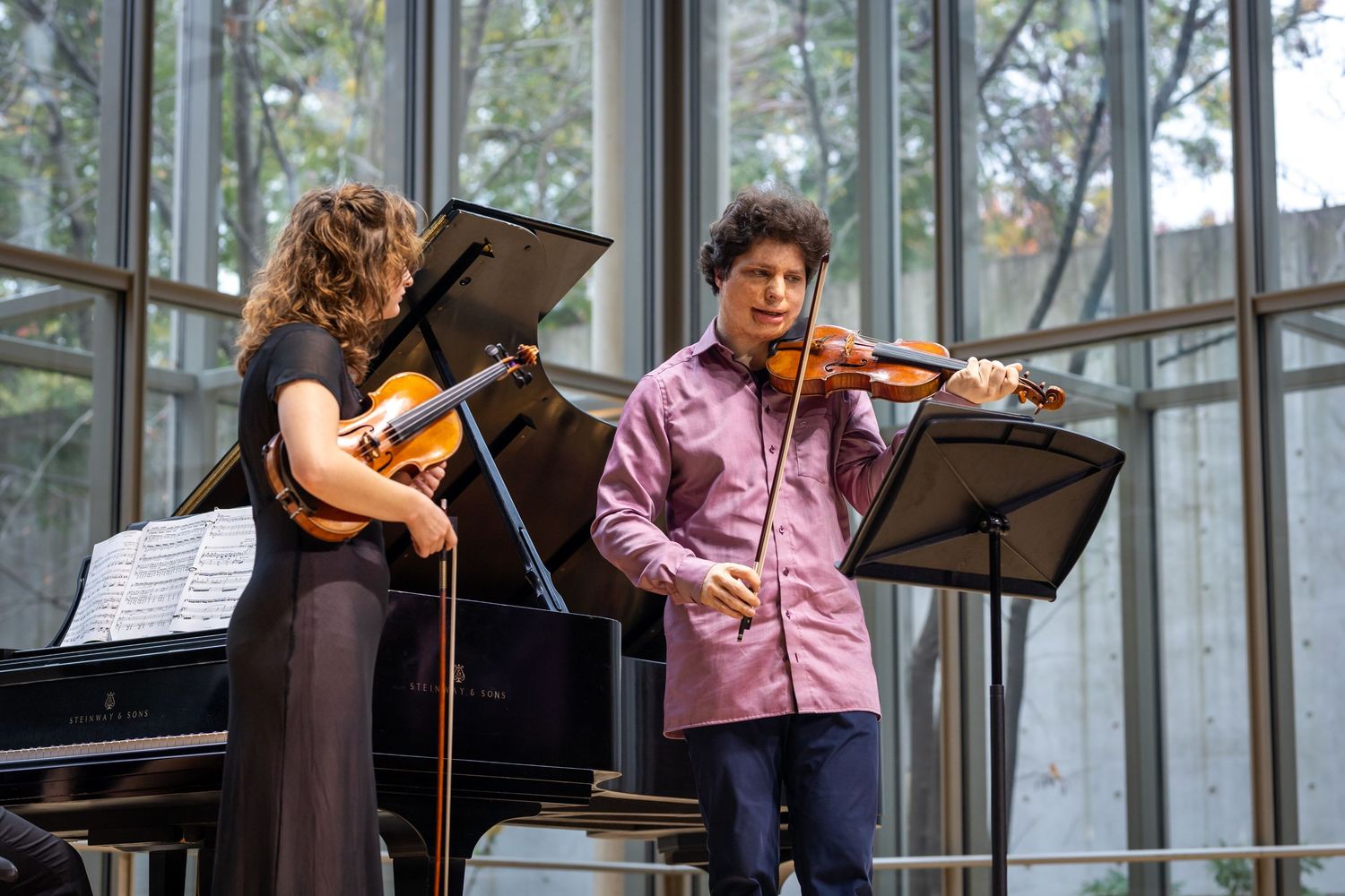 Two musicians perform a violin and piano duet in a modern concert hall with floor-to-ceiling windows.