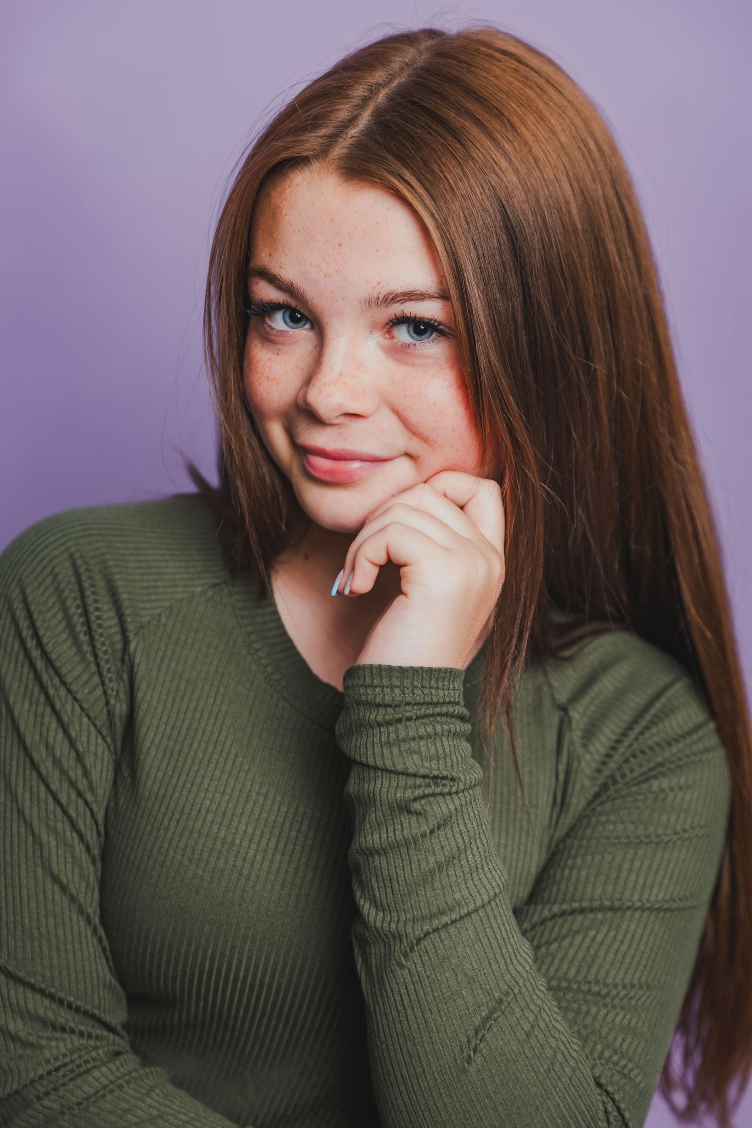Teen girl with long red hair and freckles in olive green top against purple background.